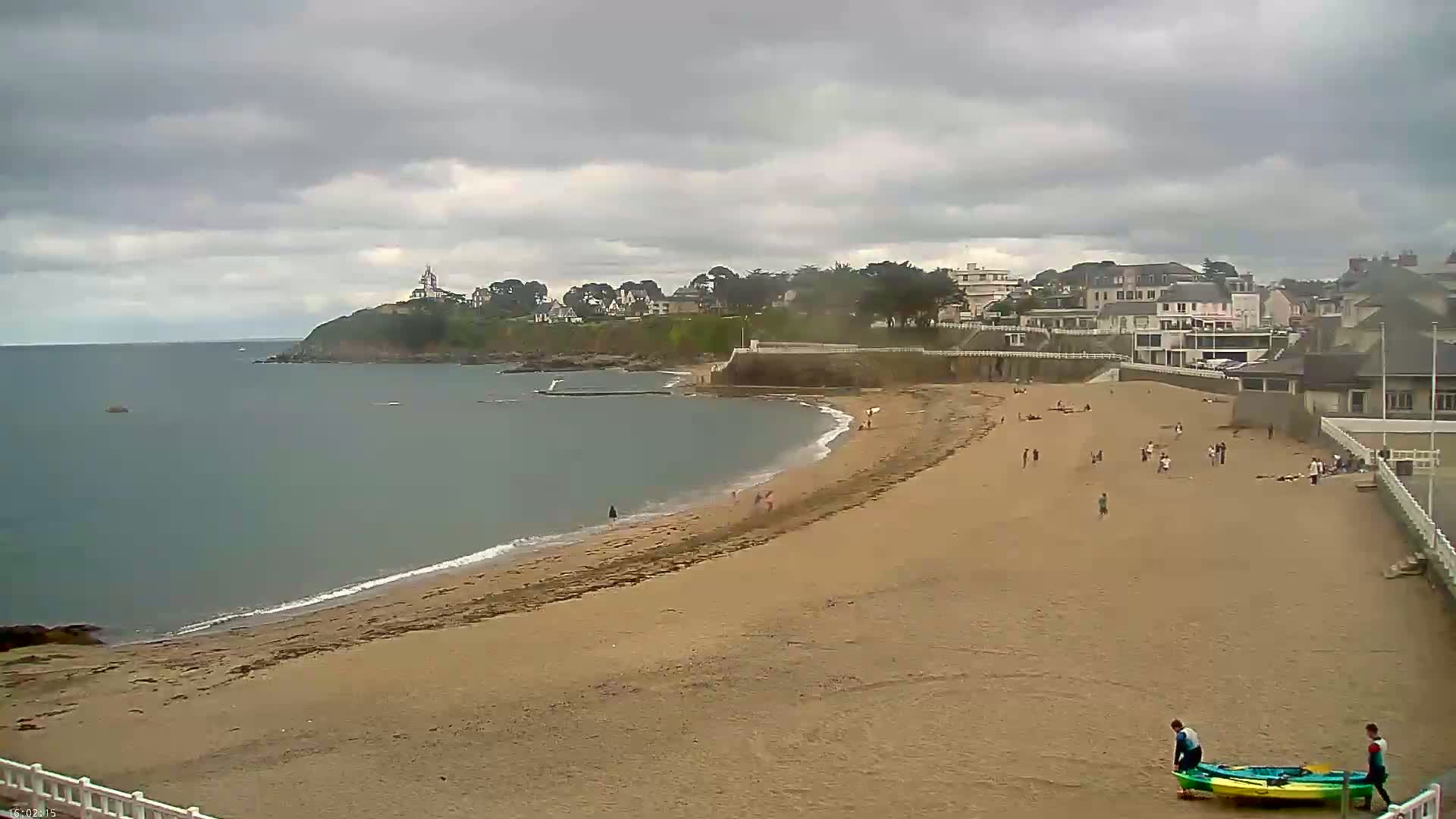 A sandy beach with a few people on it curves around a calm bay toward a town under a cloudy sky.