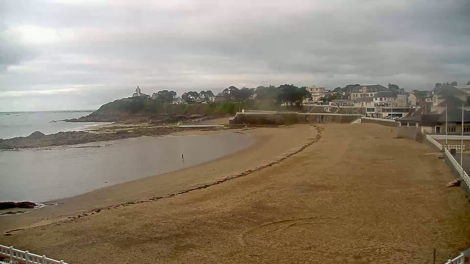 A sandy beach curves along a calm body of water under an overcast sky, with buildings and a small hill visible in the background.