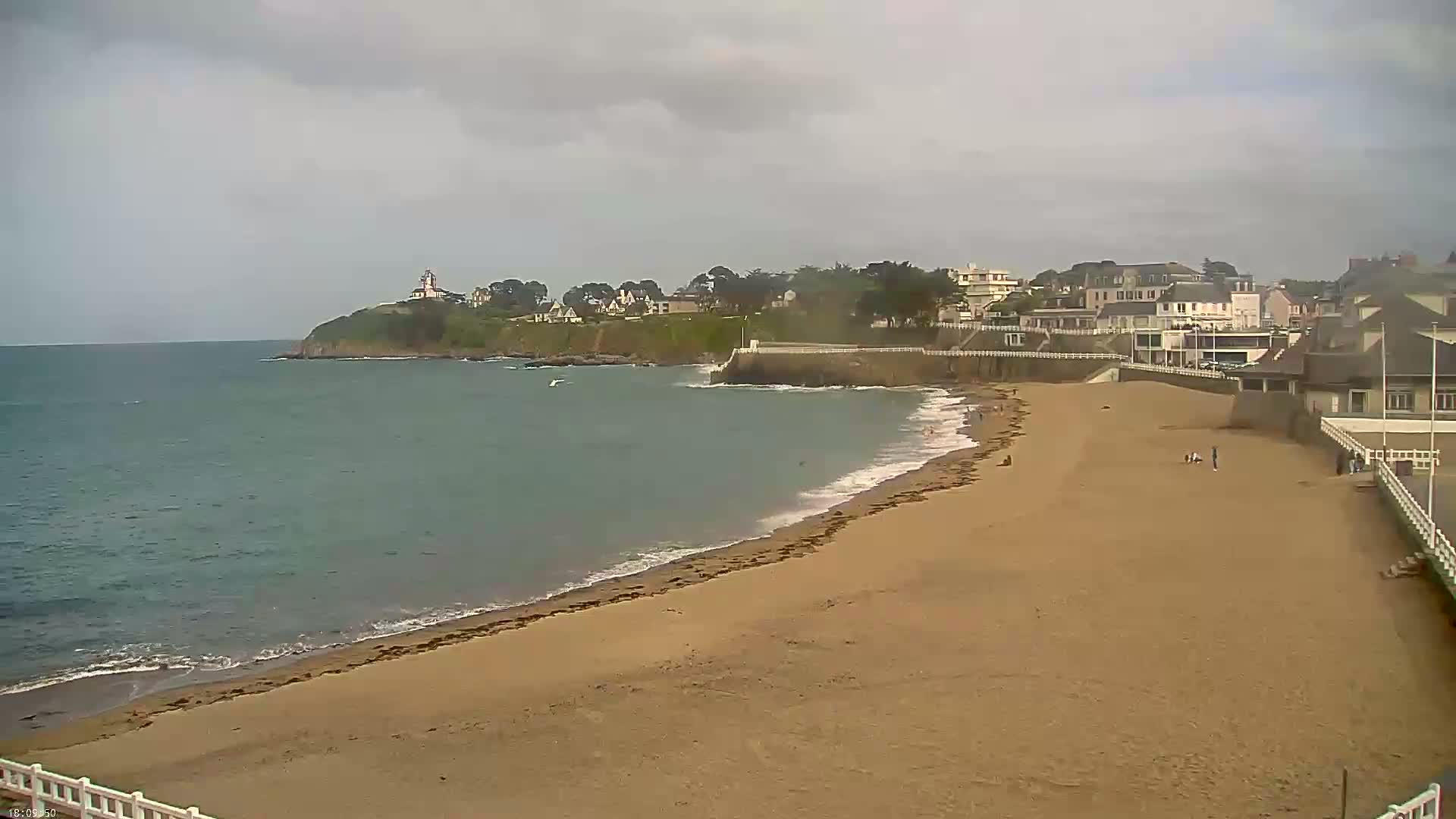 A sandy beach stretches from a calm, gray ocean toward a coastline featuring buildings and a lighthouse under an overcast sky.