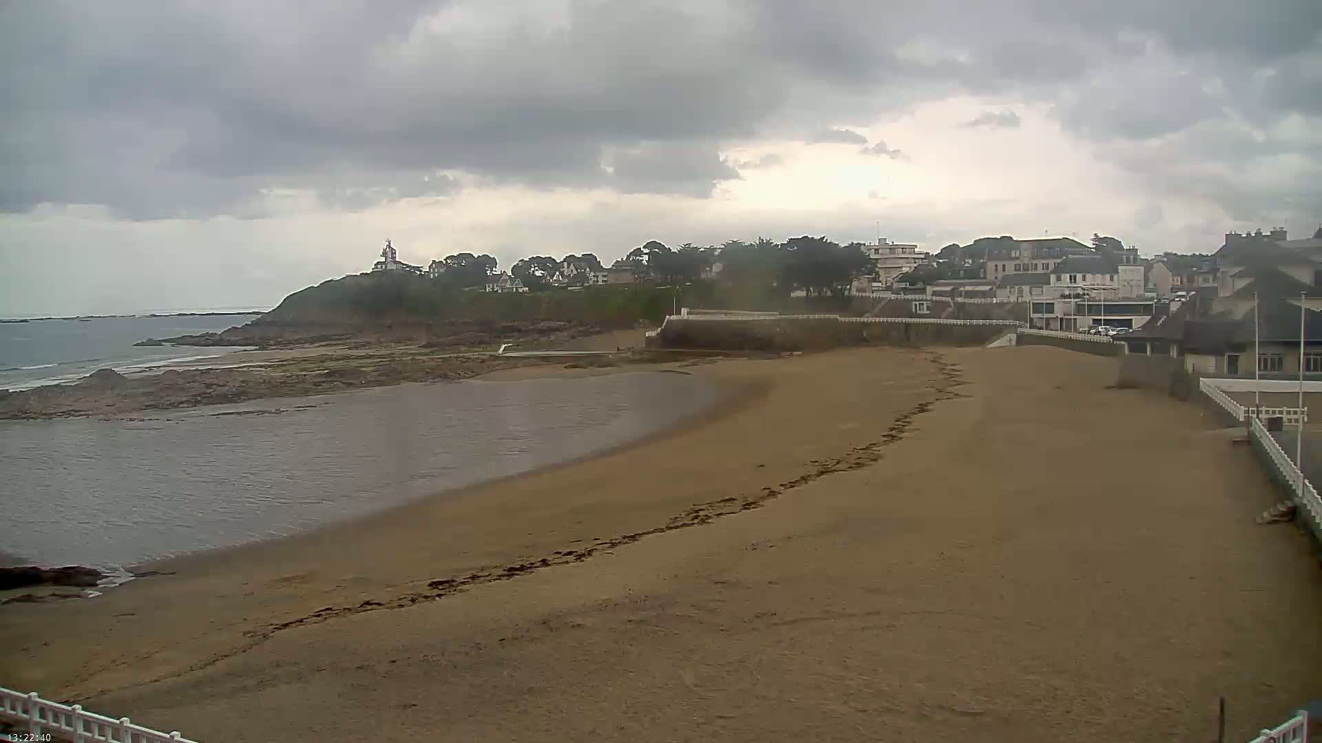 A sandy beach curves around a calm bay, under an overcast sky, with a small town visible on a nearby hill.