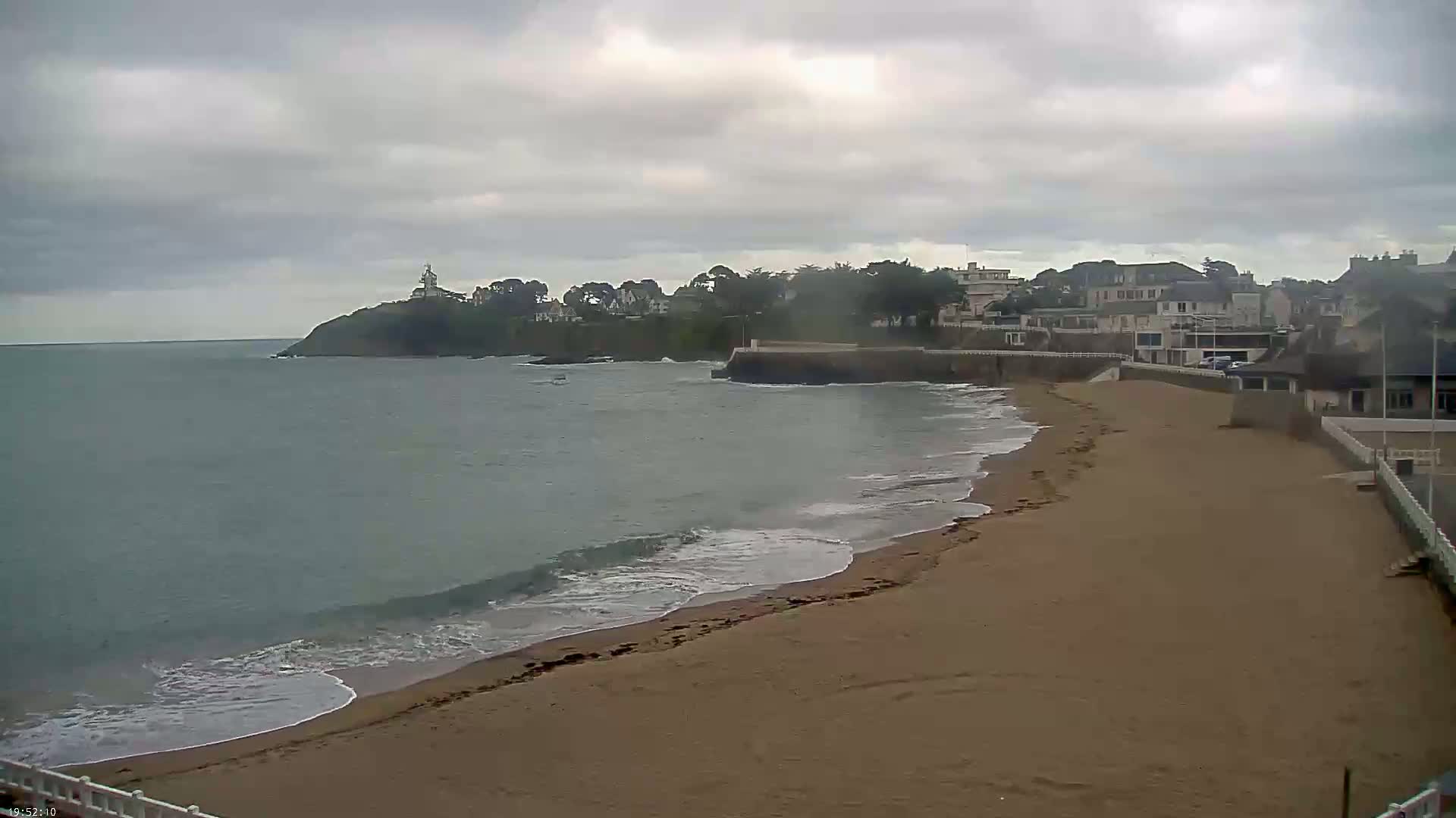 A sandy beach with gentle waves laps the shore under a cloudy sky, next to a line of houses and a small, rocky point jutting into the sea.