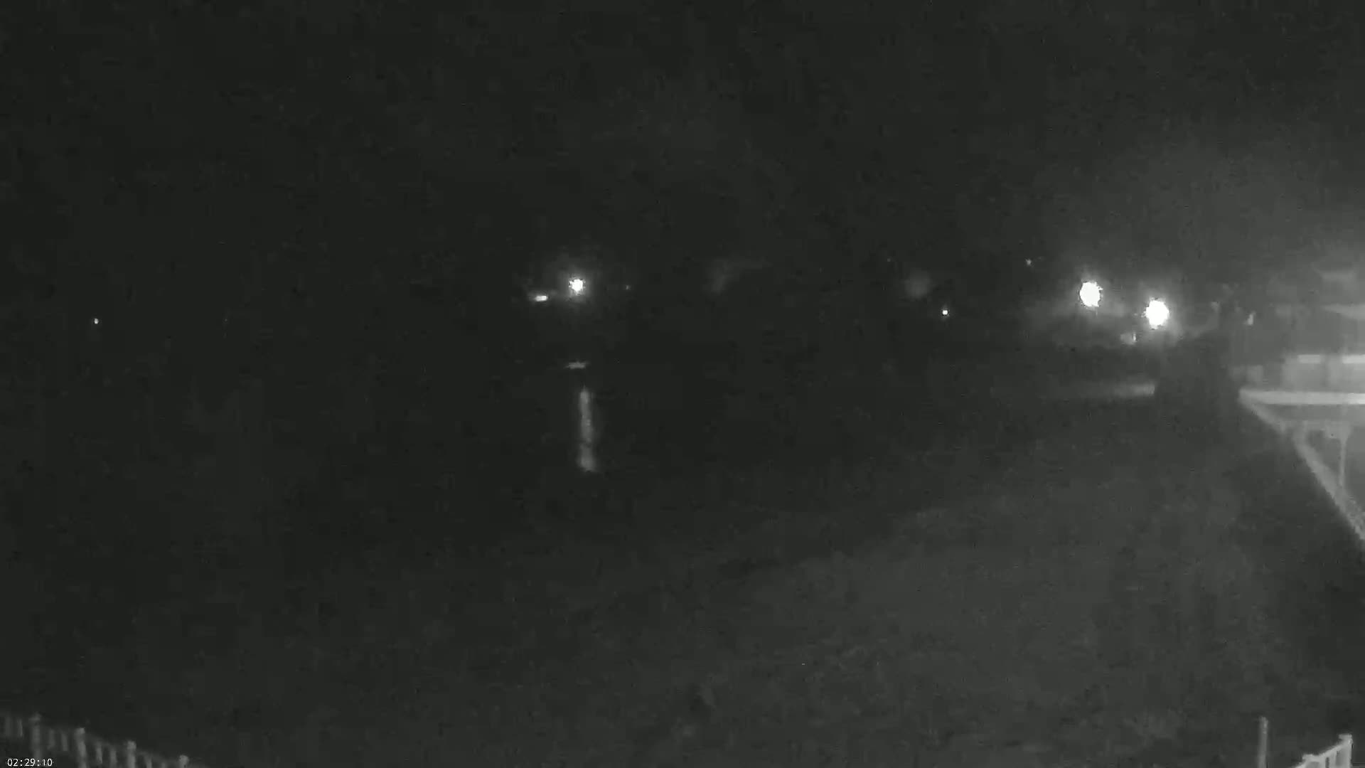 A nighttime, low-light image shows a dark body of water, possibly a lake or river, with faint lights visible in the distance and a portion of a deck or structure in the lower right corner.