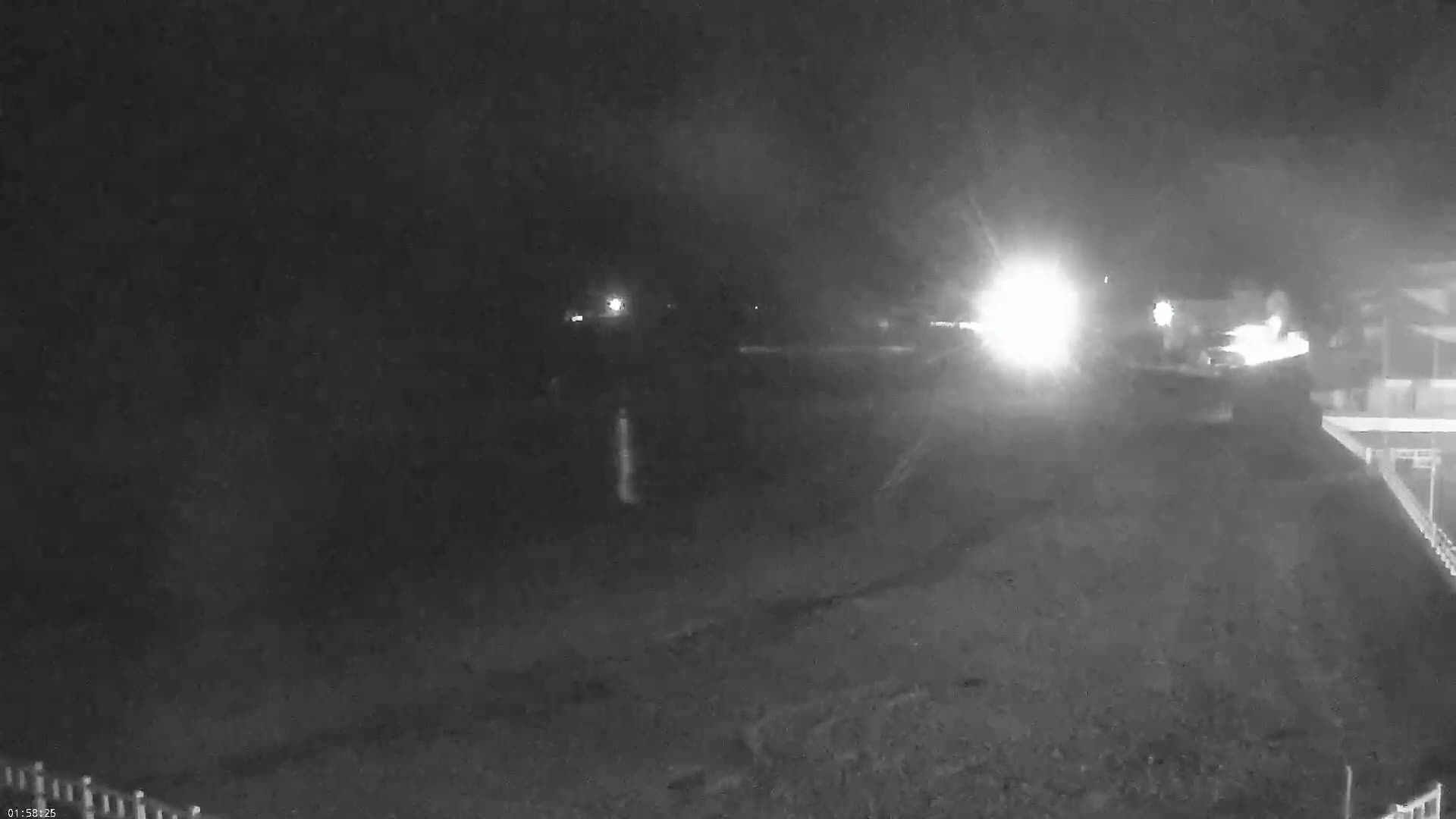 A nighttime, hazy, low-light view of a dark, empty beach area with some distant lights and a building's edge visible in the bottom right.
