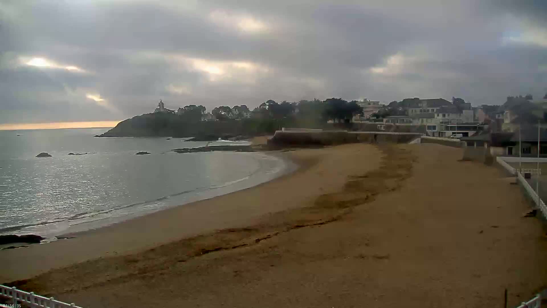 A sandy beach curves gently into a calm sea under an overcast sky, with houses and a lighthouse visible on a headland in the distance.