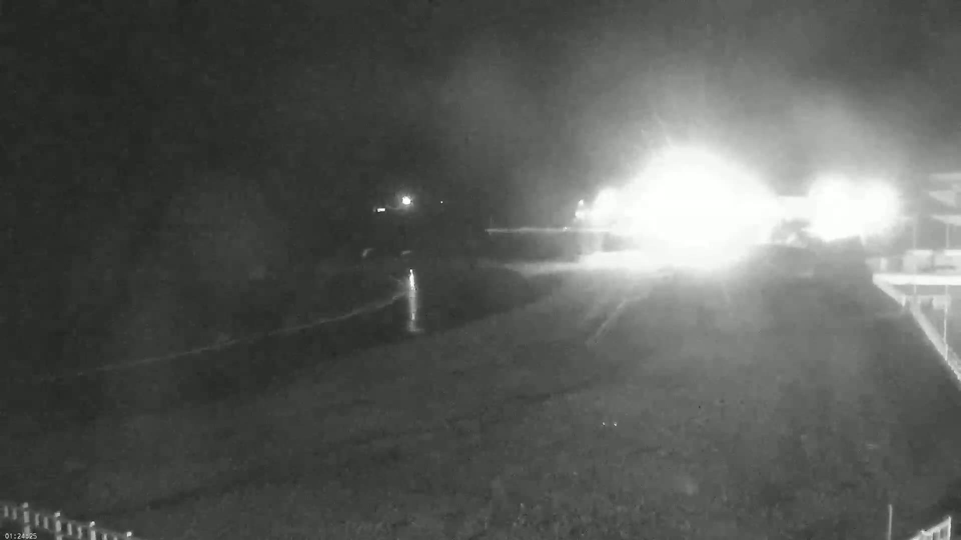 A nighttime, low-light view shows a wet, dark road with bright headlights approaching from the middle distance in heavy rain.