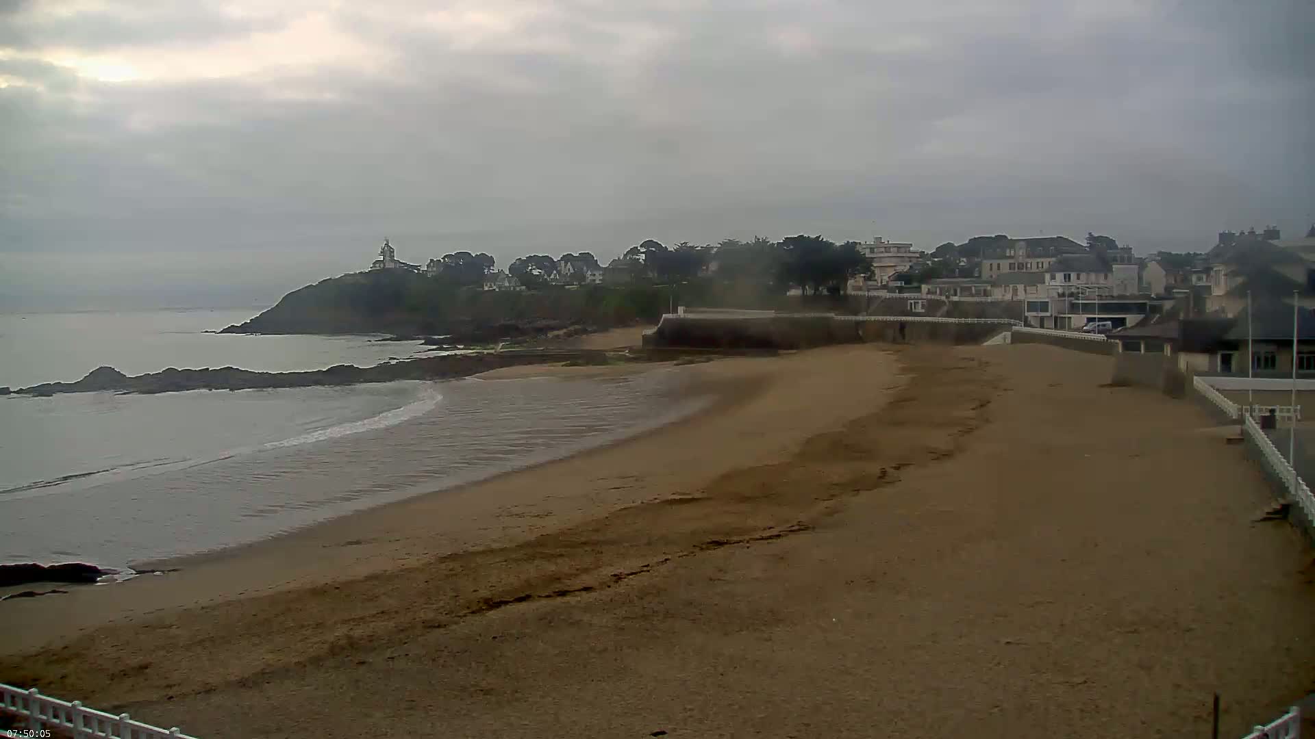 A sandy beach curves gently around a rocky point, where a small town sits under a cloudy sky.