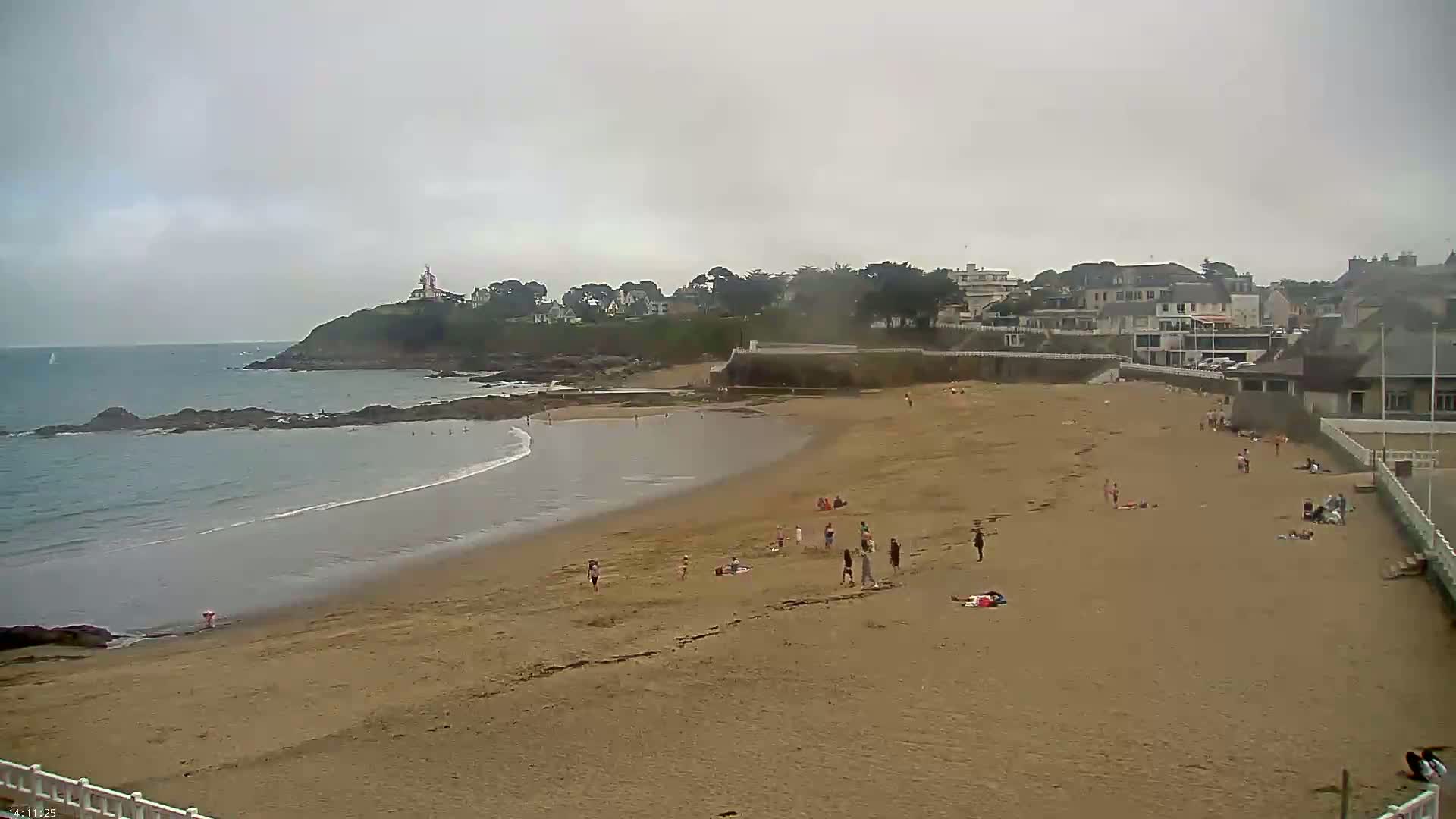 A sandy beach with a few people on it, overlooks a calm ocean under an overcast sky, and a small town sits on a hill in the background.