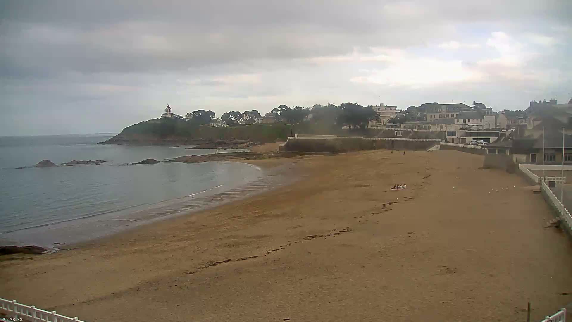 A sandy beach curves around a calm bay under an overcast sky, with a small town and a lighthouse visible in the distance.