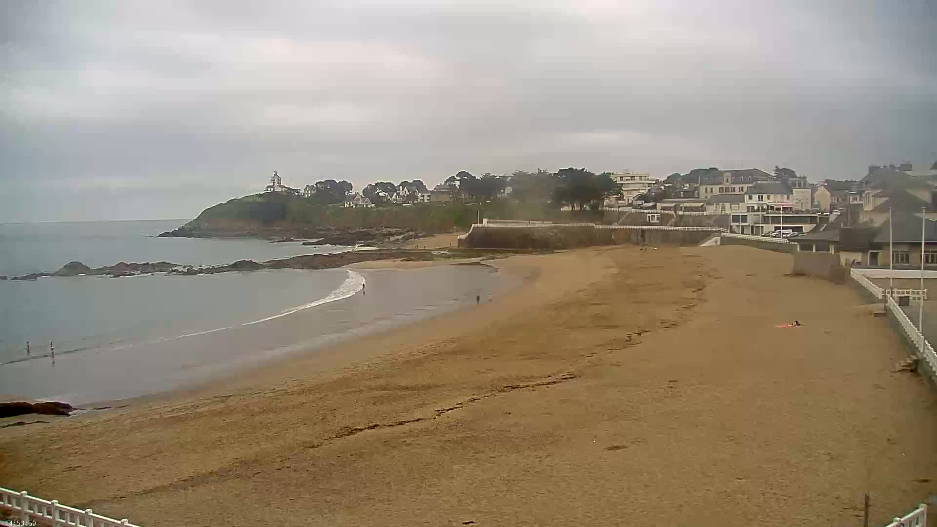 A sandy beach curves gently around a rocky point, with calm waves lapping the shore under an overcast sky, and a small town visible beyond the beach.