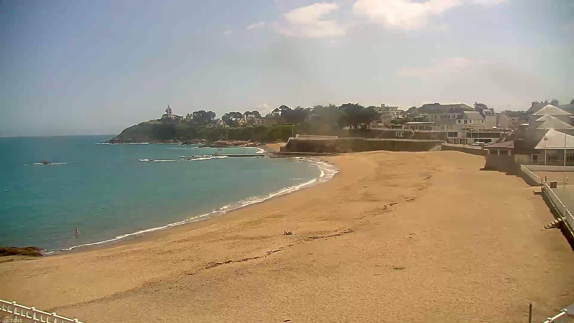 A sandy beach curves along a calm, teal ocean, with a small town and a lighthouse visible on a cliff in the background under a mostly sunny sky.