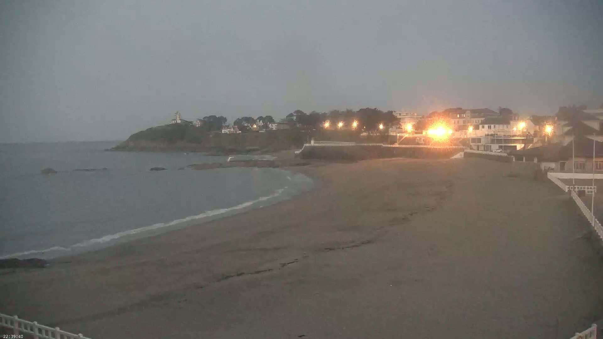 A sandy beach at dusk or early evening, gently lapped by waves under a cloudy sky, faces a coastline with buildings and a lighthouse.