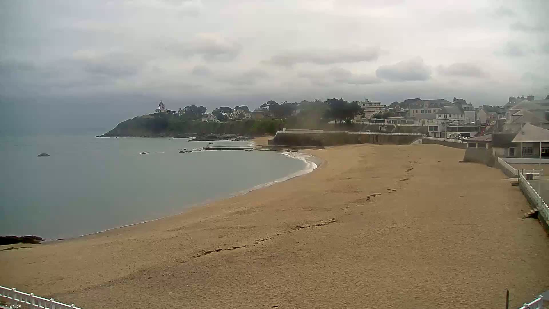 A sandy beach curves along a calm, gray sea under an overcast sky, with buildings and a lighthouse visible on a hill in the distance.