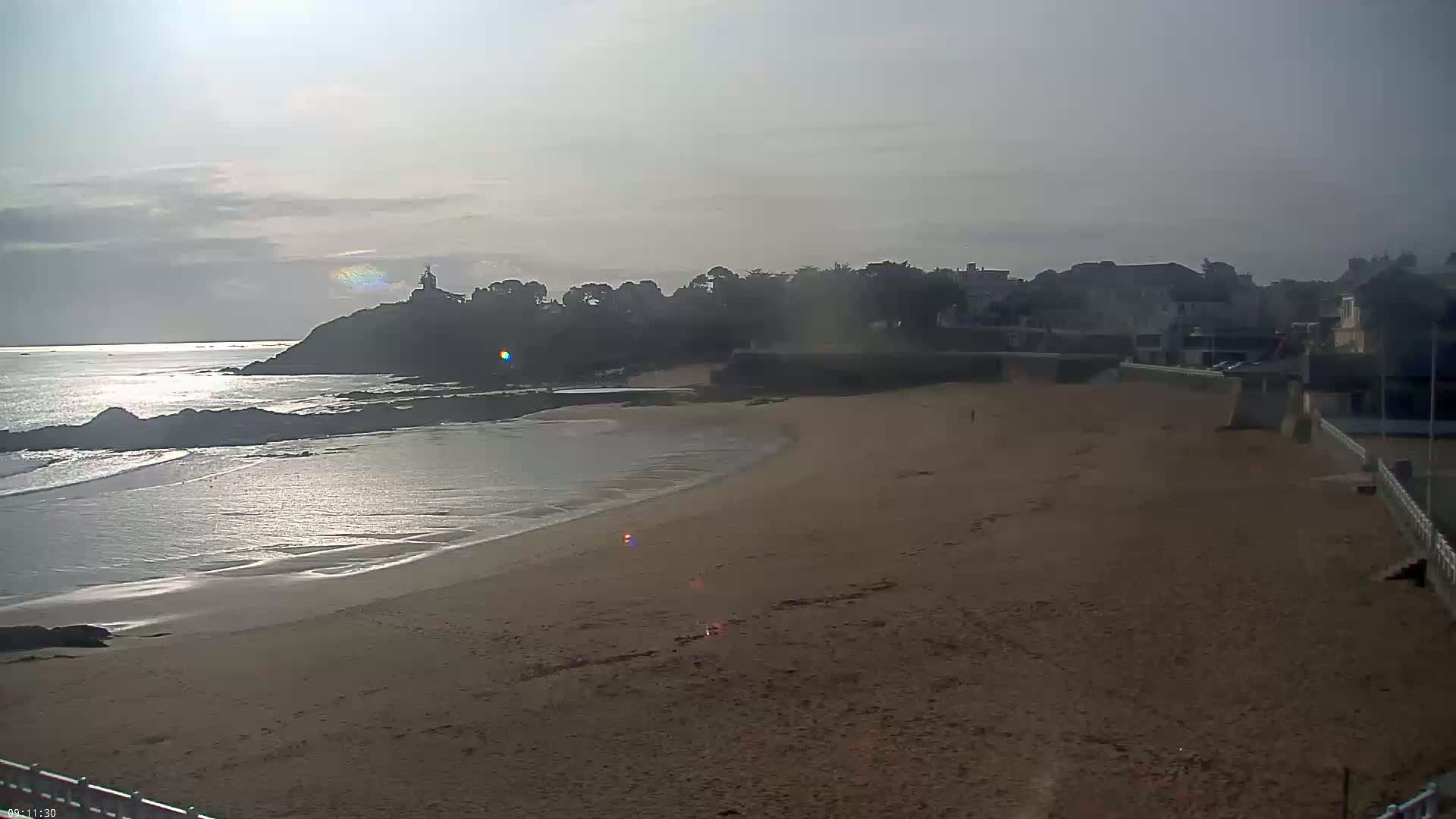 A mostly sunny day shows a sandy beach curving around a rocky point, where a small town sits on the cliff overlooking the calm ocean.