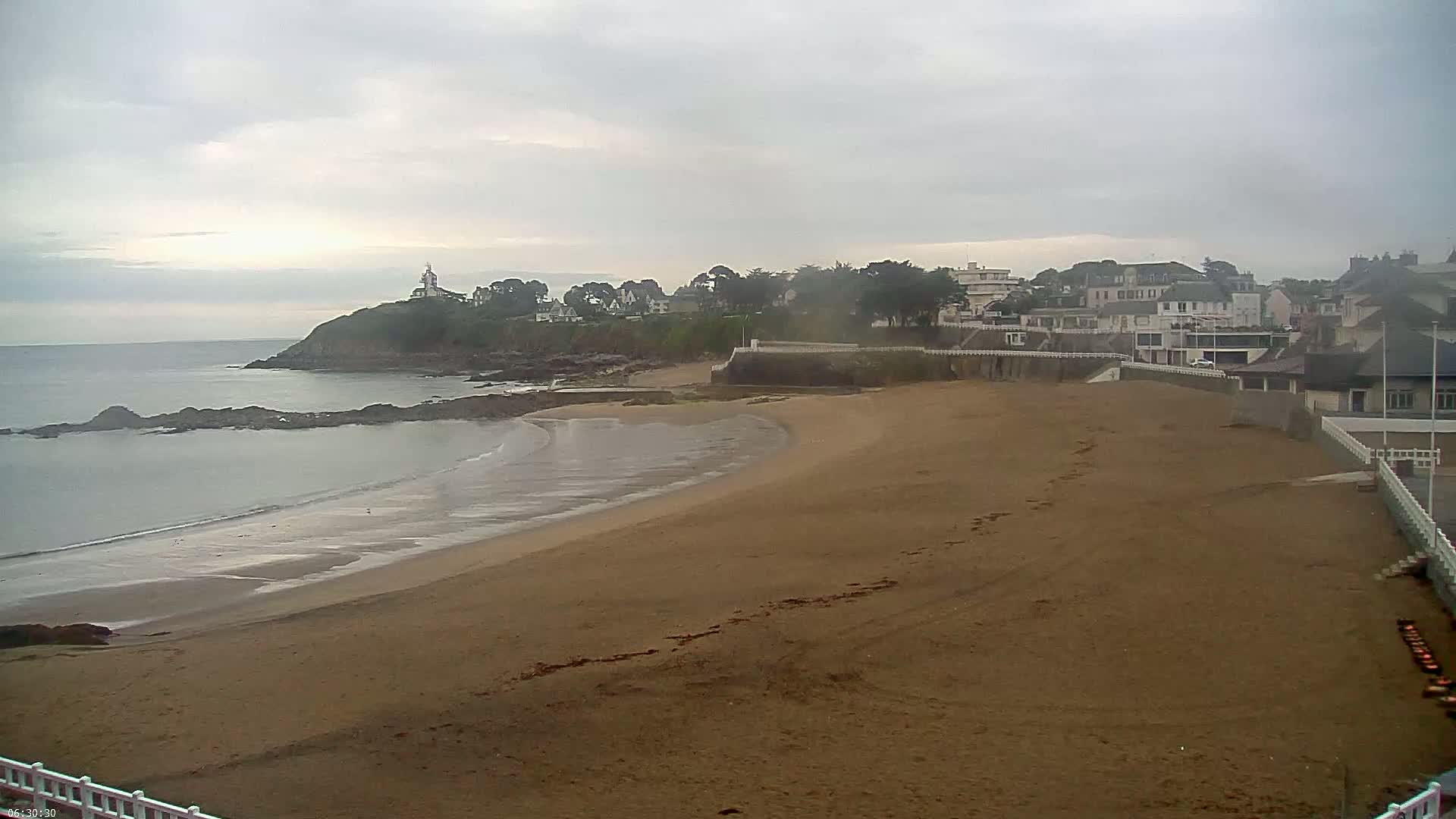 A sandy beach curves around a calm bay under an overcast sky, with a village and a lighthouse visible in the distance.