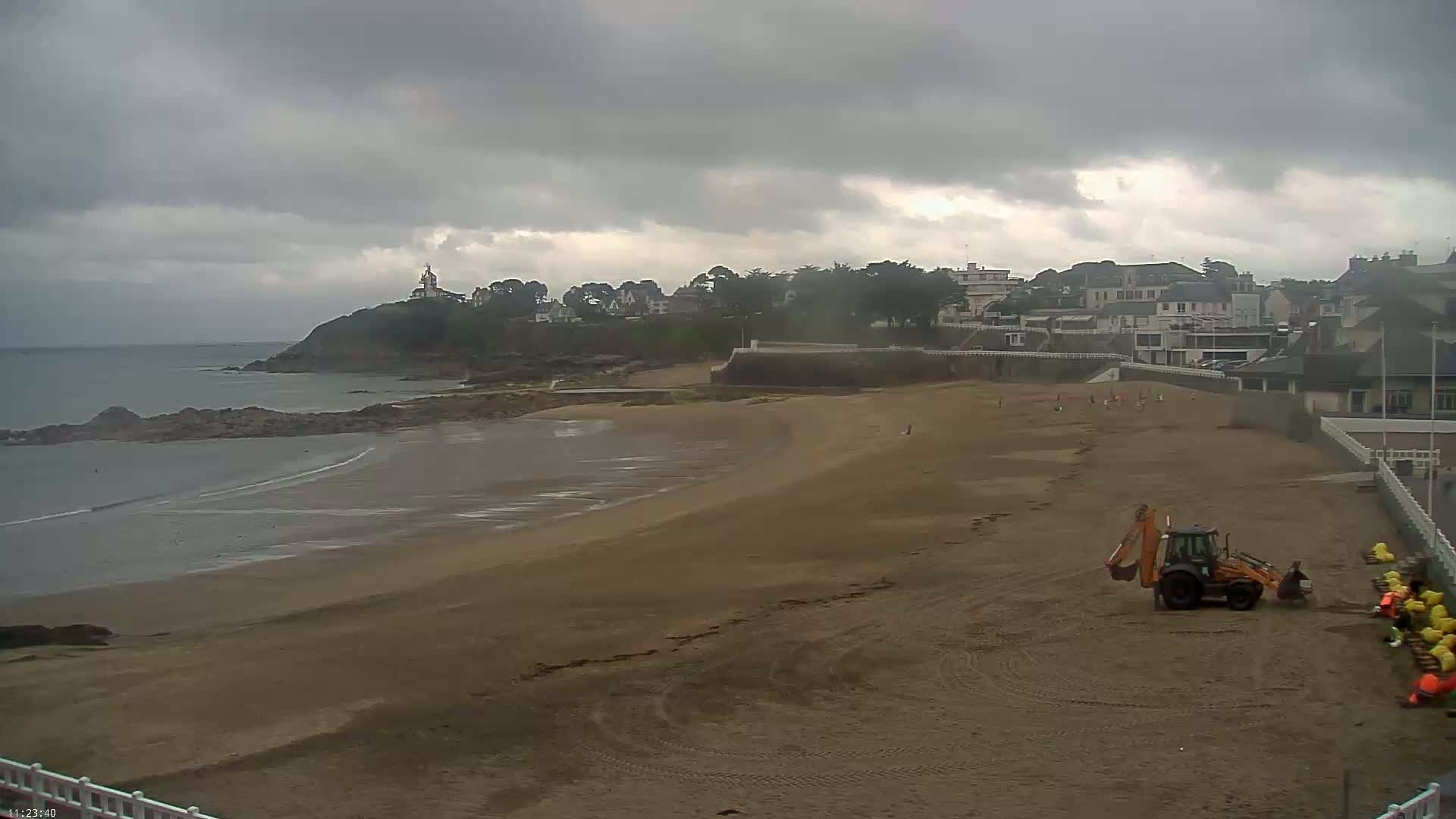 A sandy beach stretches out under an overcast sky, with a small town visible in the background and a backhoe working near some yellow objects on the sand.