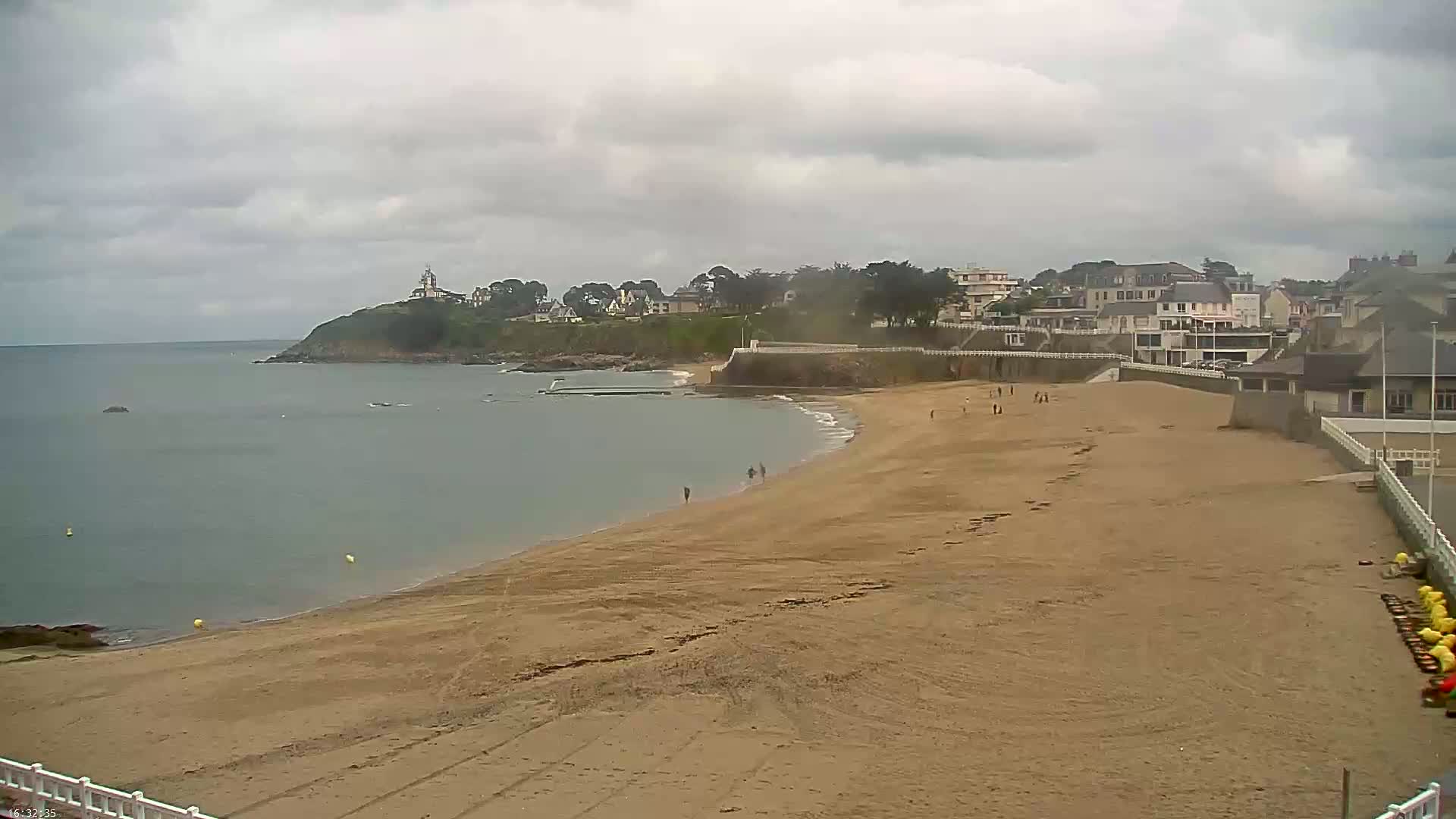 A sandy beach stretches along a calm, gray sea under an overcast sky, with a small town and a hilltop building visible in the distance.