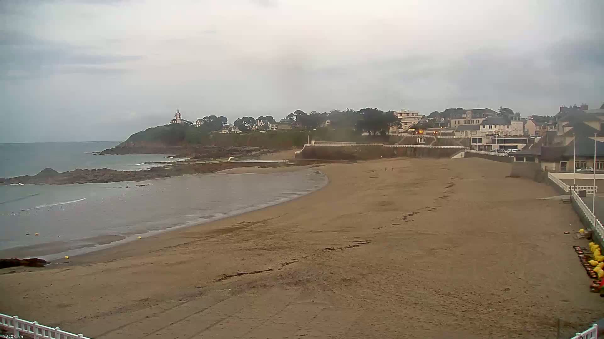 A sandy beach curves gently toward a calm ocean under an overcast sky, with buildings and a small lighthouse visible on a cliff in the distance.