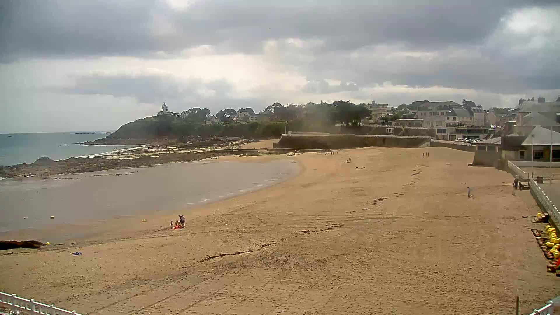 A sandy beach stretches below a town nestled against a rocky coastline under a cloudy sky.