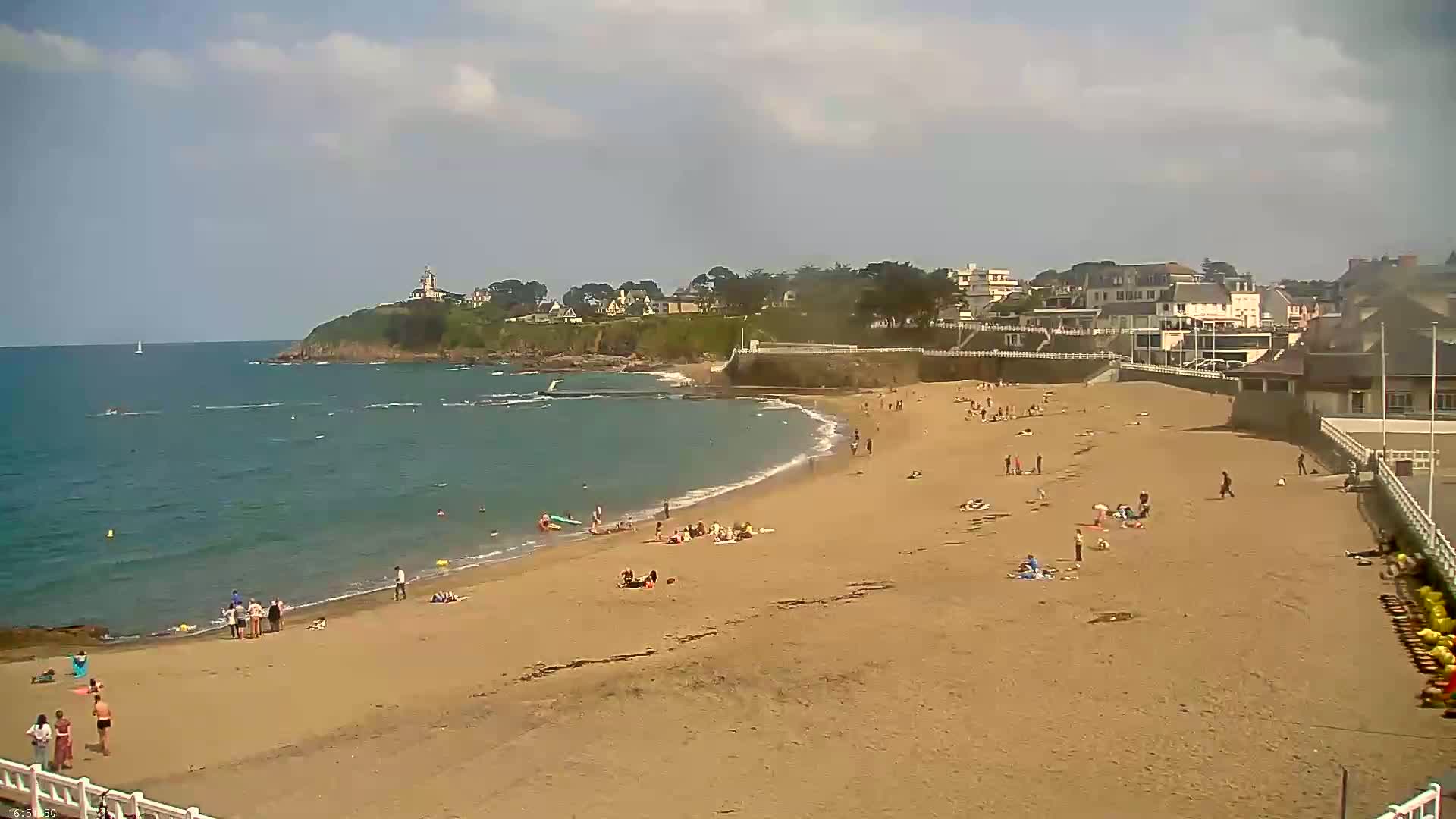 A sandy beach with scattered sunbathers is bordered by calm ocean water and a hillside town under a mostly sunny sky.