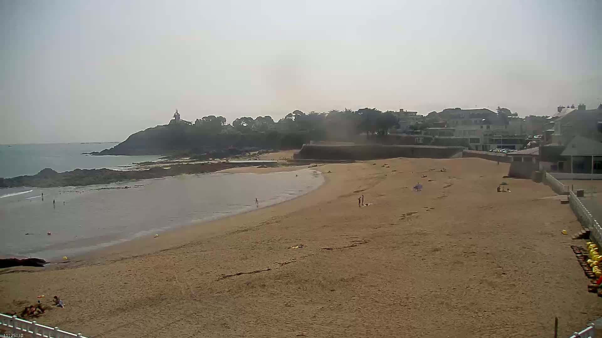 A sandy beach with a few people on it curves gently around a calm bay, bordered by buildings and a rocky headland under a hazy sky.