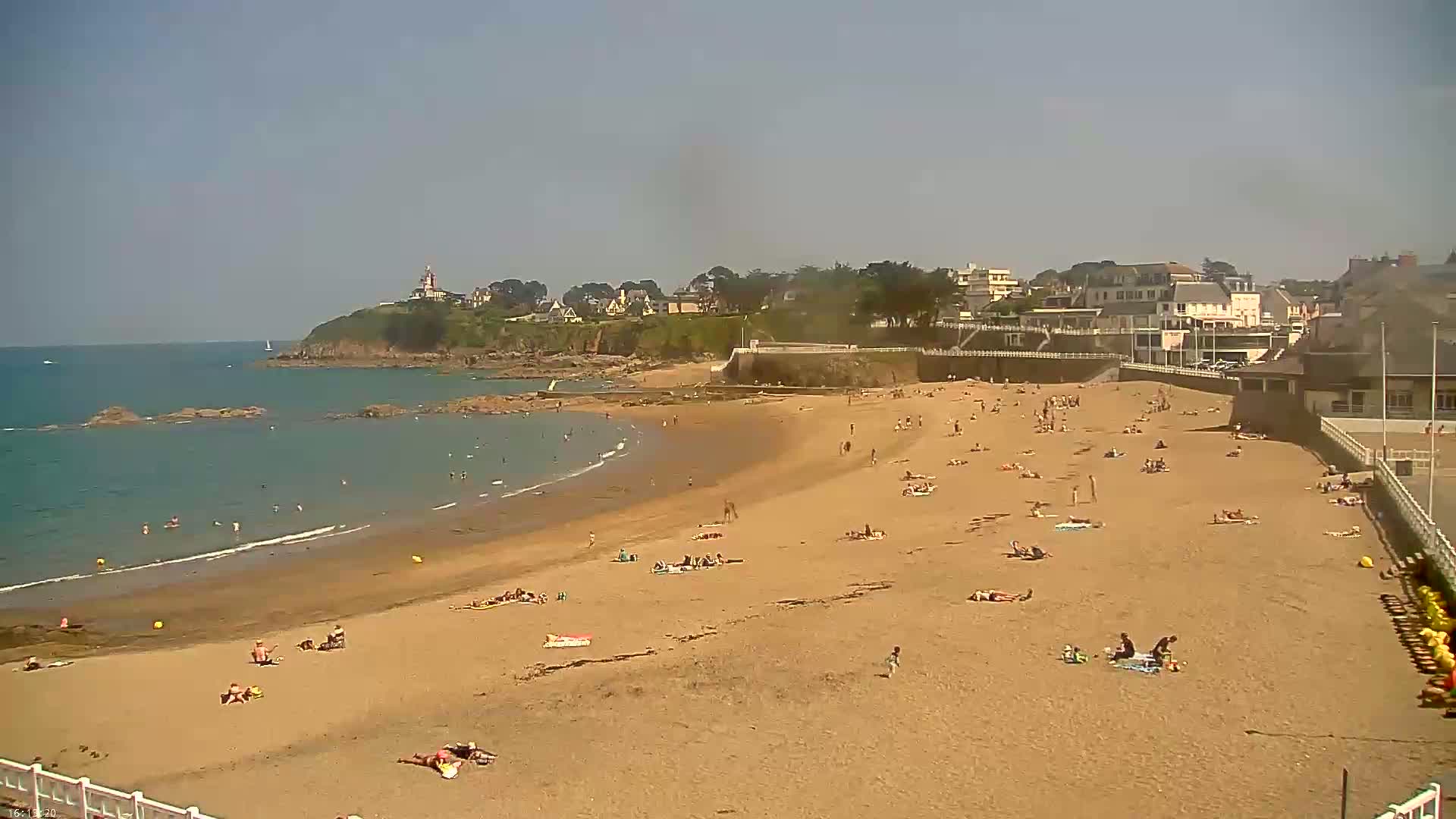 A sandy beach with many people sunbathing and swimming, backed by a seaside town under a clear, sunny sky.