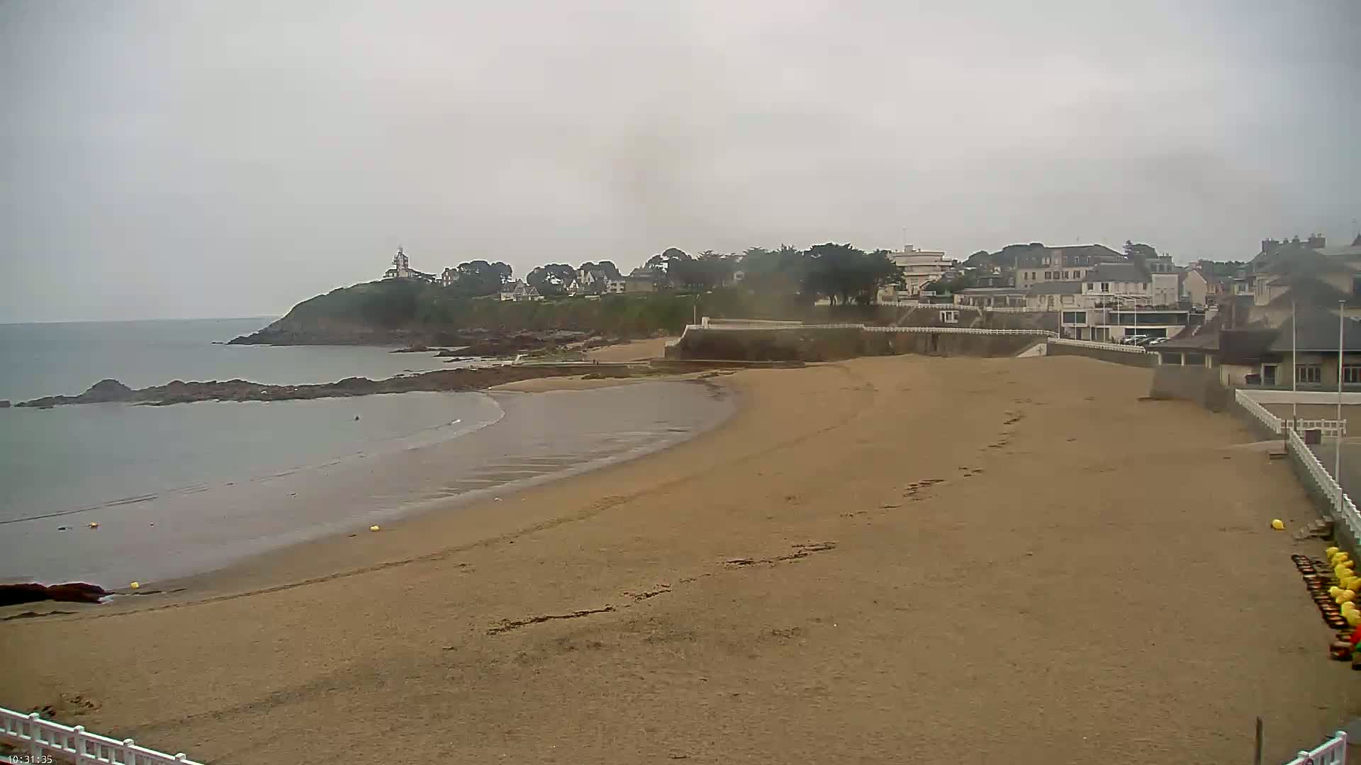 A sandy beach curves around a rocky point, with calm, grey water, buildings, and greenery visible in the background under an overcast sky.