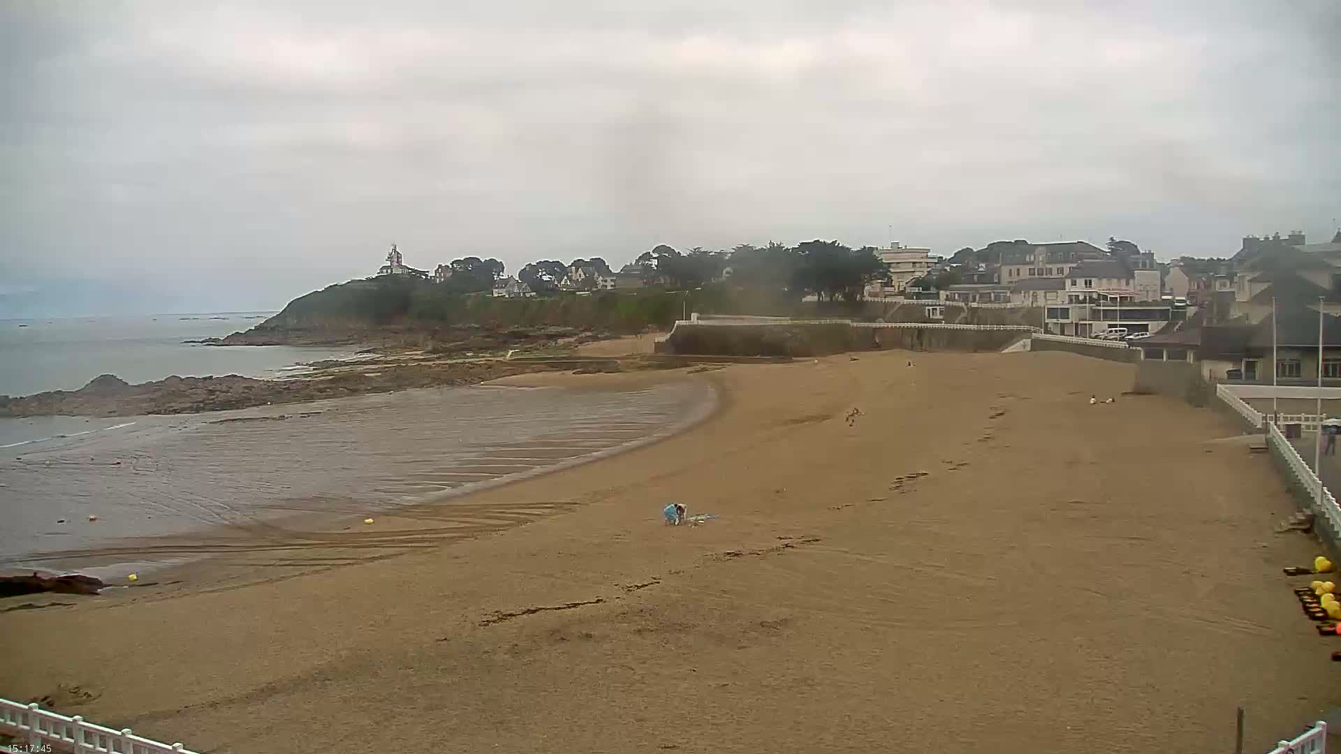 A mostly empty sandy beach curves around a rocky point, with a calm, overcast sky and a few buildings visible in the distance.