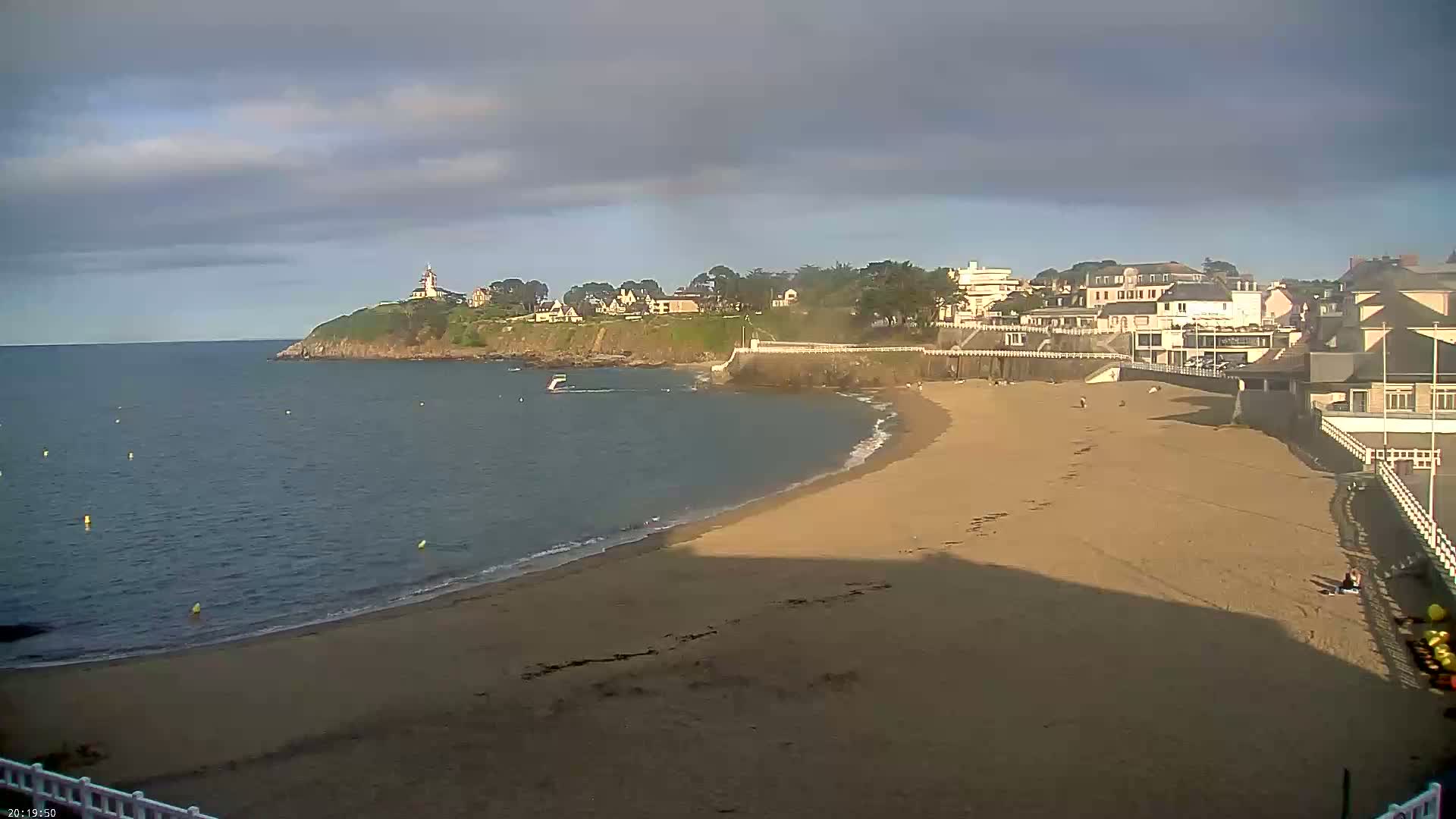 A sandy beach curves along a calm ocean, with buildings and a lighthouse visible on a nearby cliff under a partly cloudy sky.