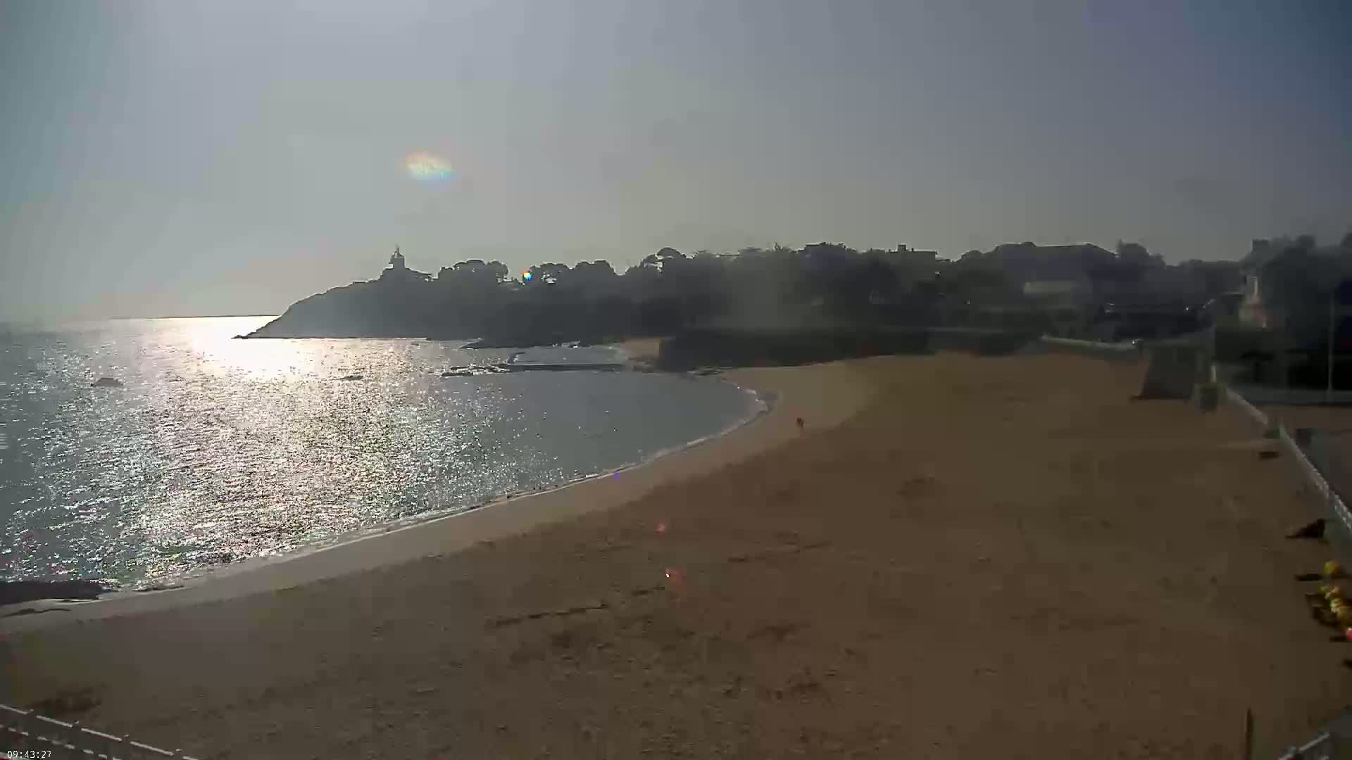 A sandy beach curves around a calm, sunlit bay, with a distant coastline featuring buildings and trees under a clear sky.