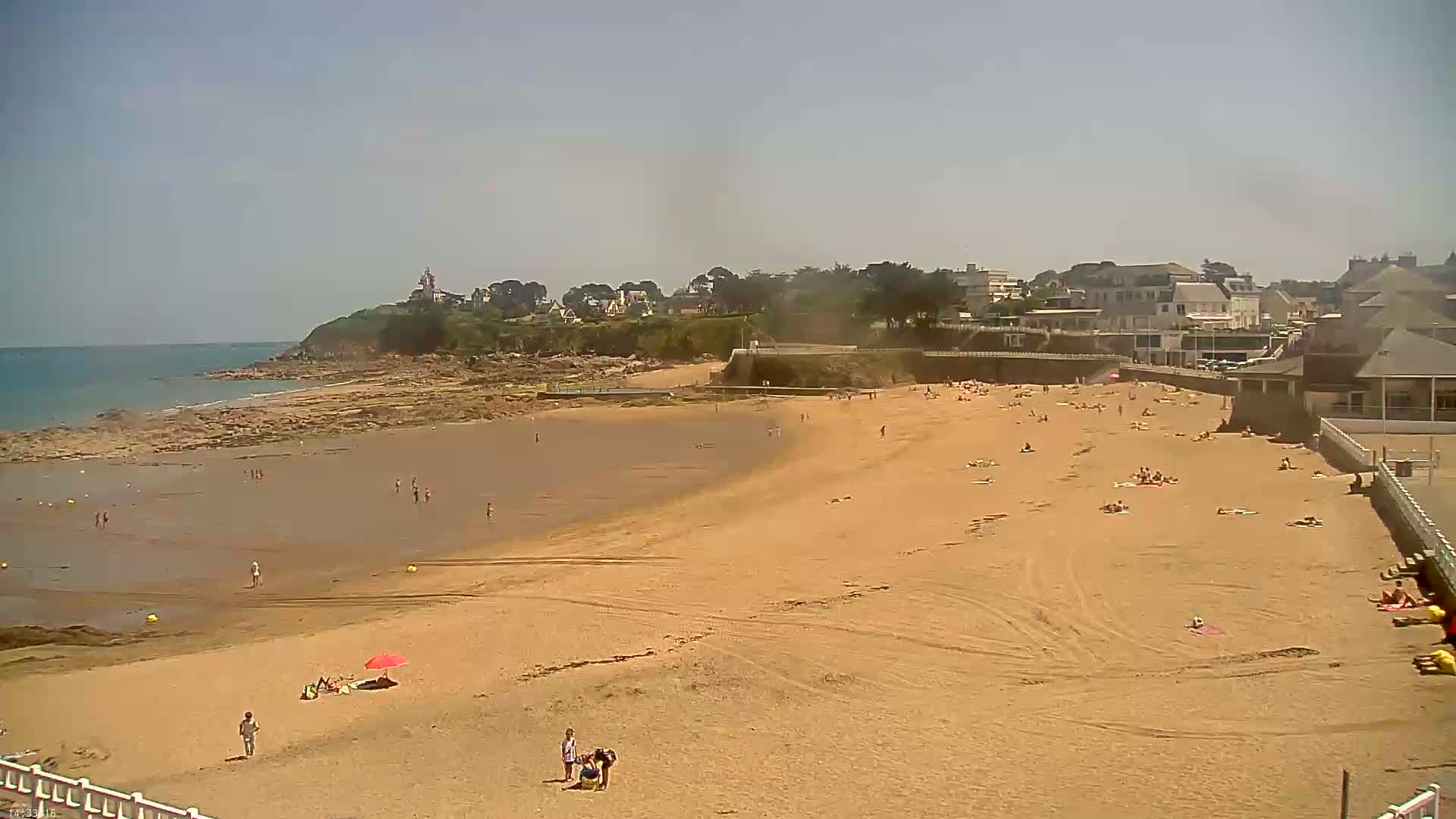 A sandy beach with scattered sunbathers stretches from a rocky shoreline to buildings and houses under a clear, sunny sky.