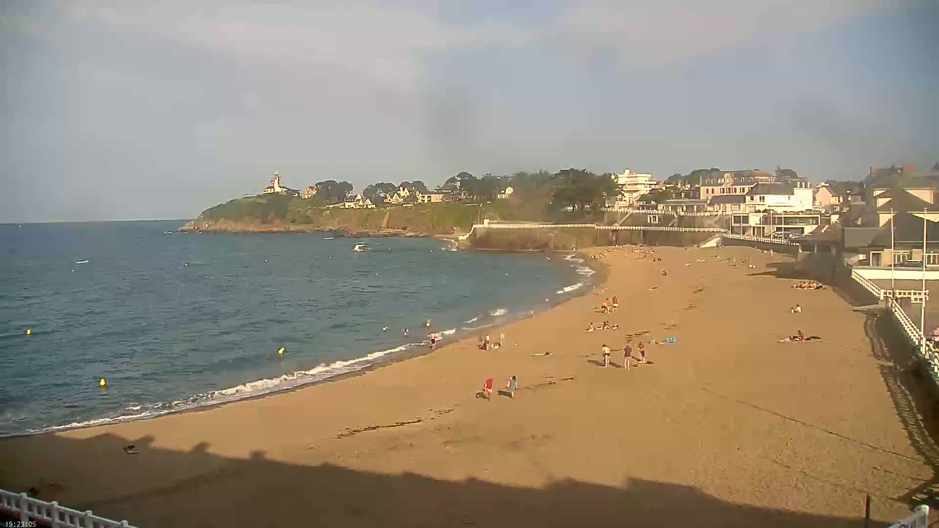 A sandy beach with a few people on it curves around a calm, blue ocean next to a coastal town on a partly sunny day.