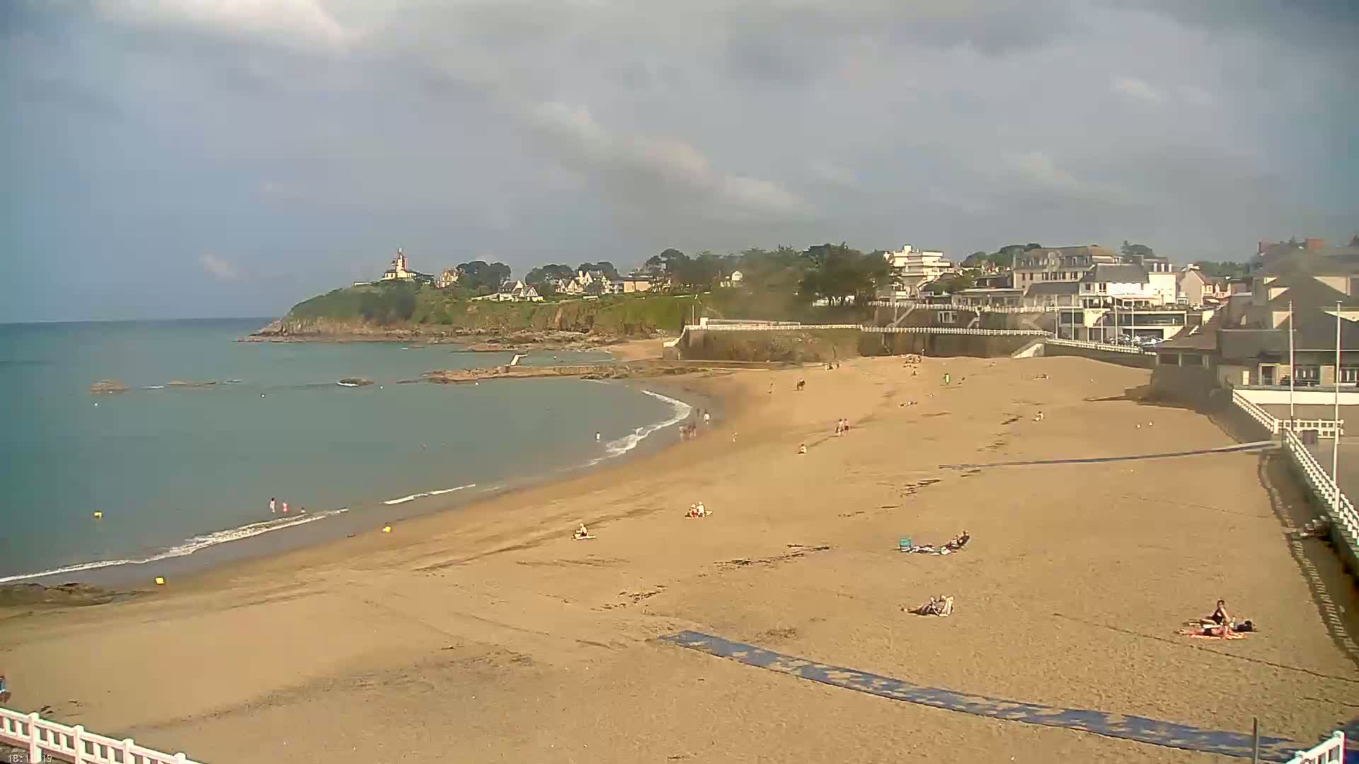 A sandy beach with a few people sunbathing and swimming stretches alongside a calm ocean under a partly cloudy sky, backed by a town and a small, rocky headland with a lighthouse.