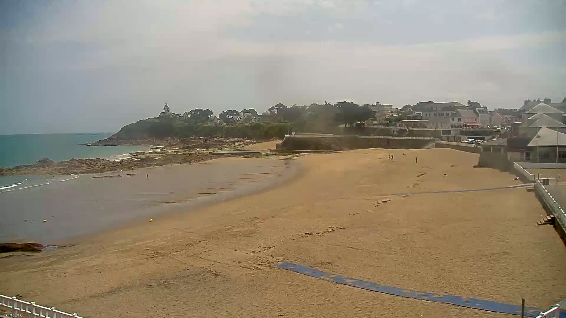 A sandy beach stretches out to a calm ocean under a mostly cloudy sky, with a small town and a lighthouse visible in the distance.