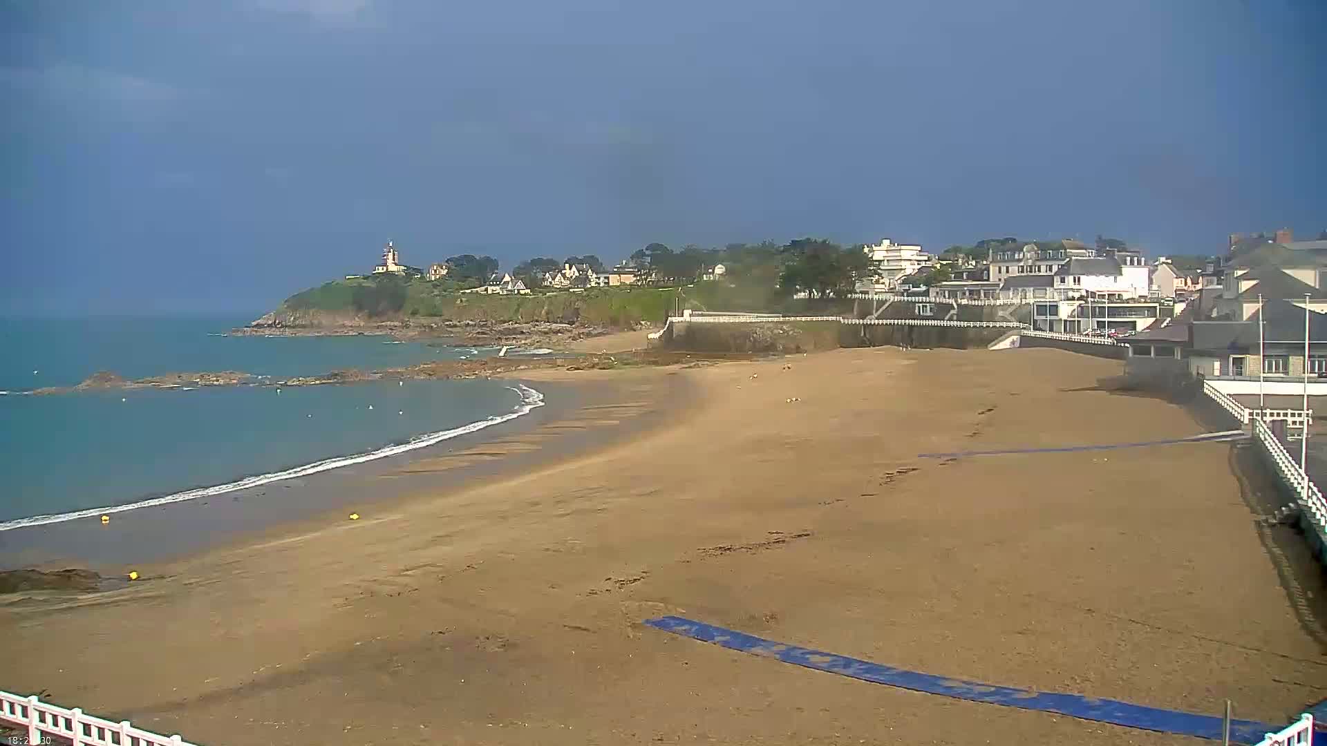 A sandy beach stretches out to a calm ocean under a mostly clear sky, with a town and a cliff in the background.