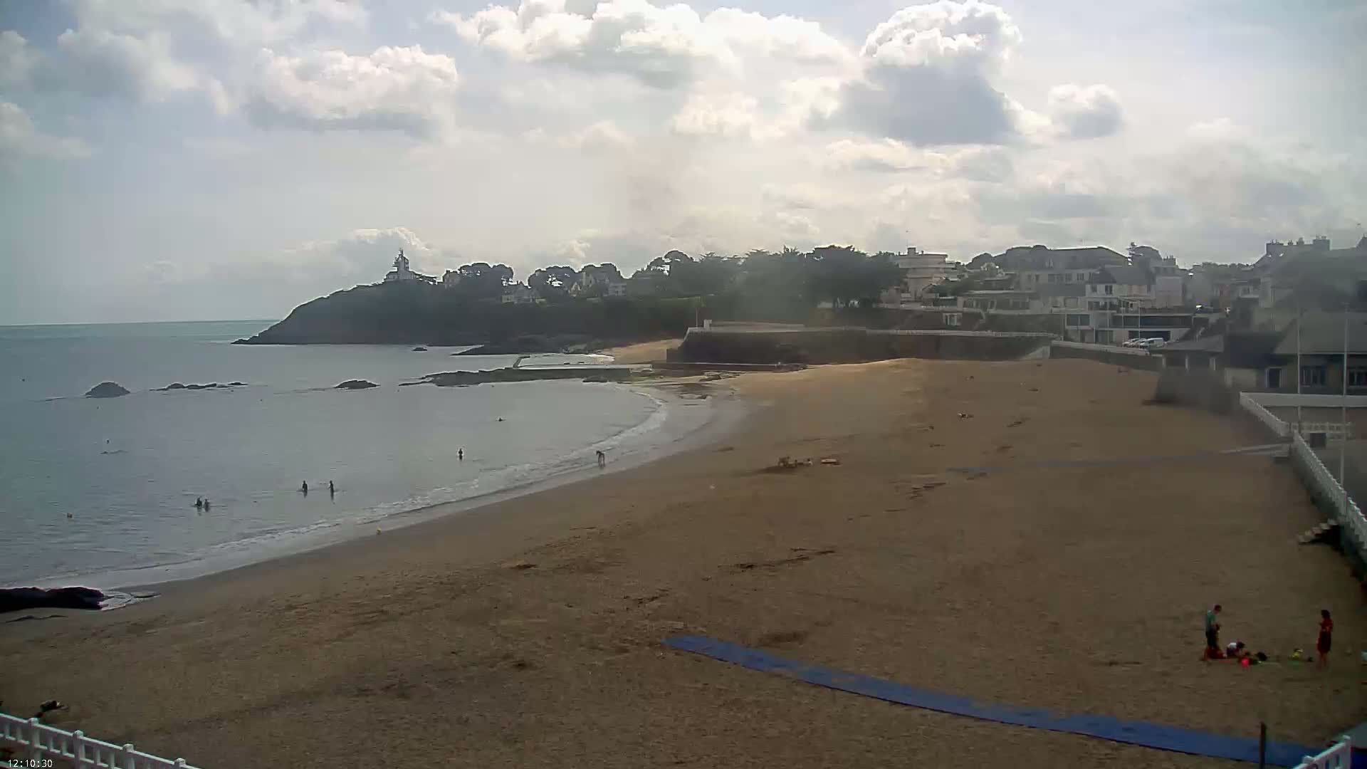 A sandy beach with a few people on it stretches out to a calm sea under a partly cloudy sky, with houses and a lighthouse visible on a nearby headland.