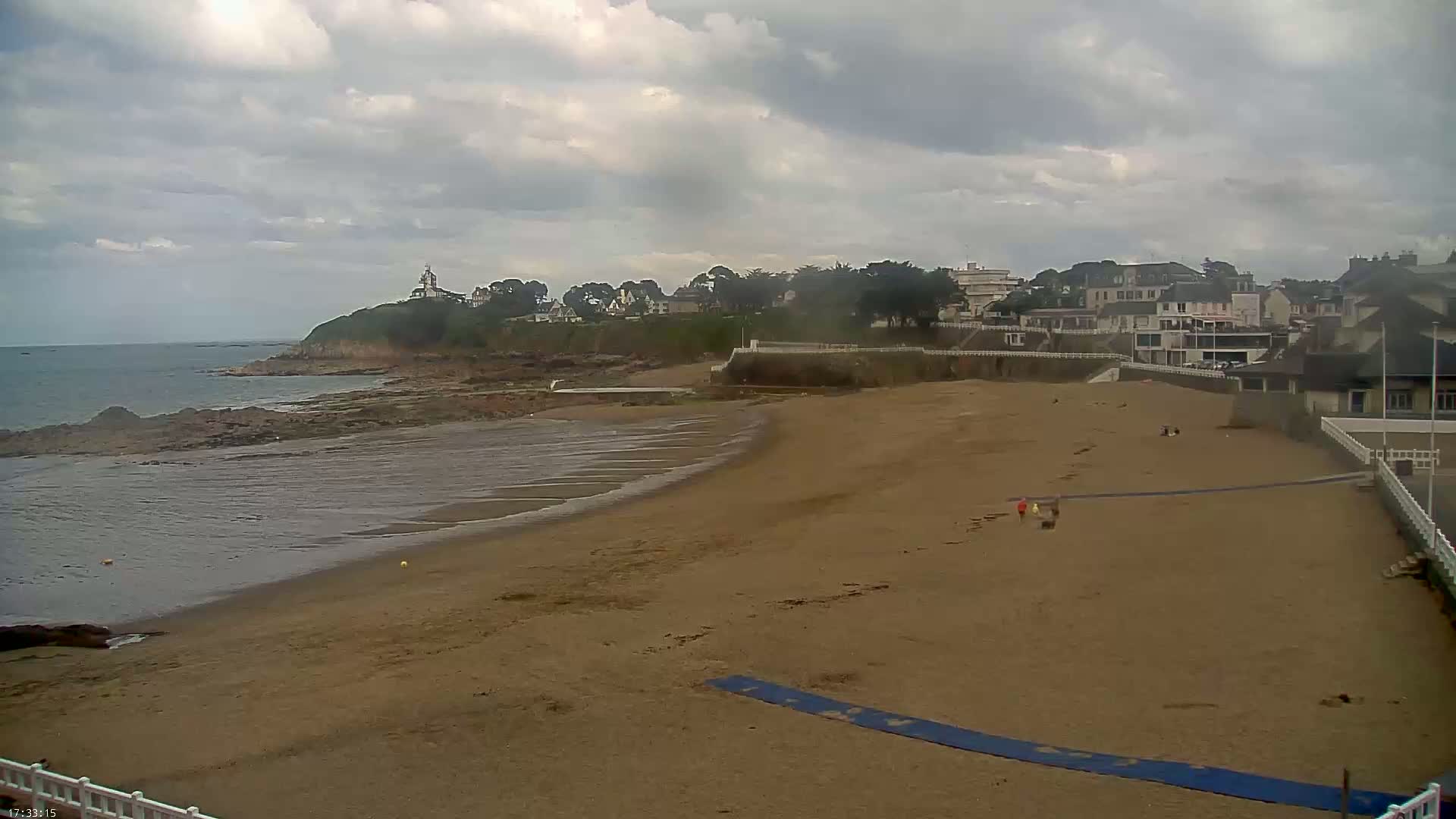A sandy beach stretches out to a calm sea under a cloudy sky, with a few people visible on the beach and buildings perched on a cliff in the background.