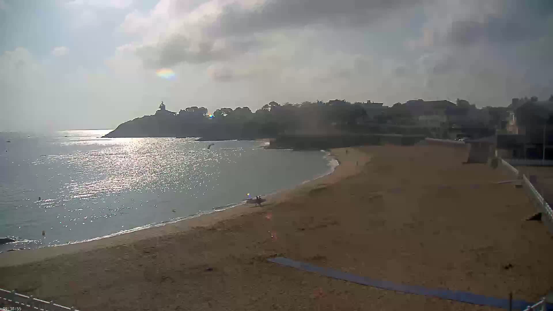 A sandy beach curves around a calm bay under a partly cloudy sky, with a distant headland featuring a structure and some buildings visible along the shore.
