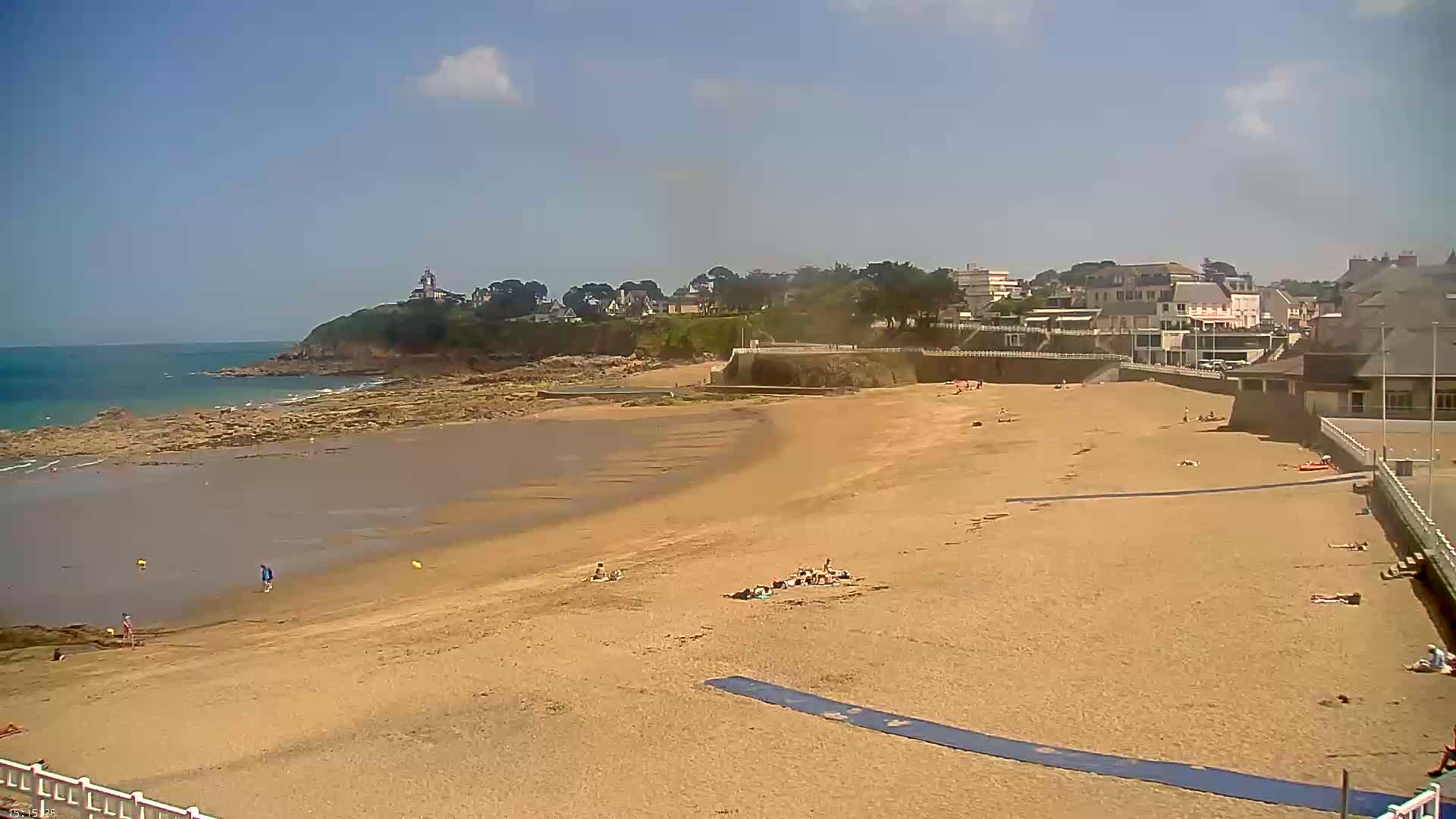 A sandy beach with scattered sunbathers stretches out to a rocky shoreline and calm ocean under a mostly sunny sky.