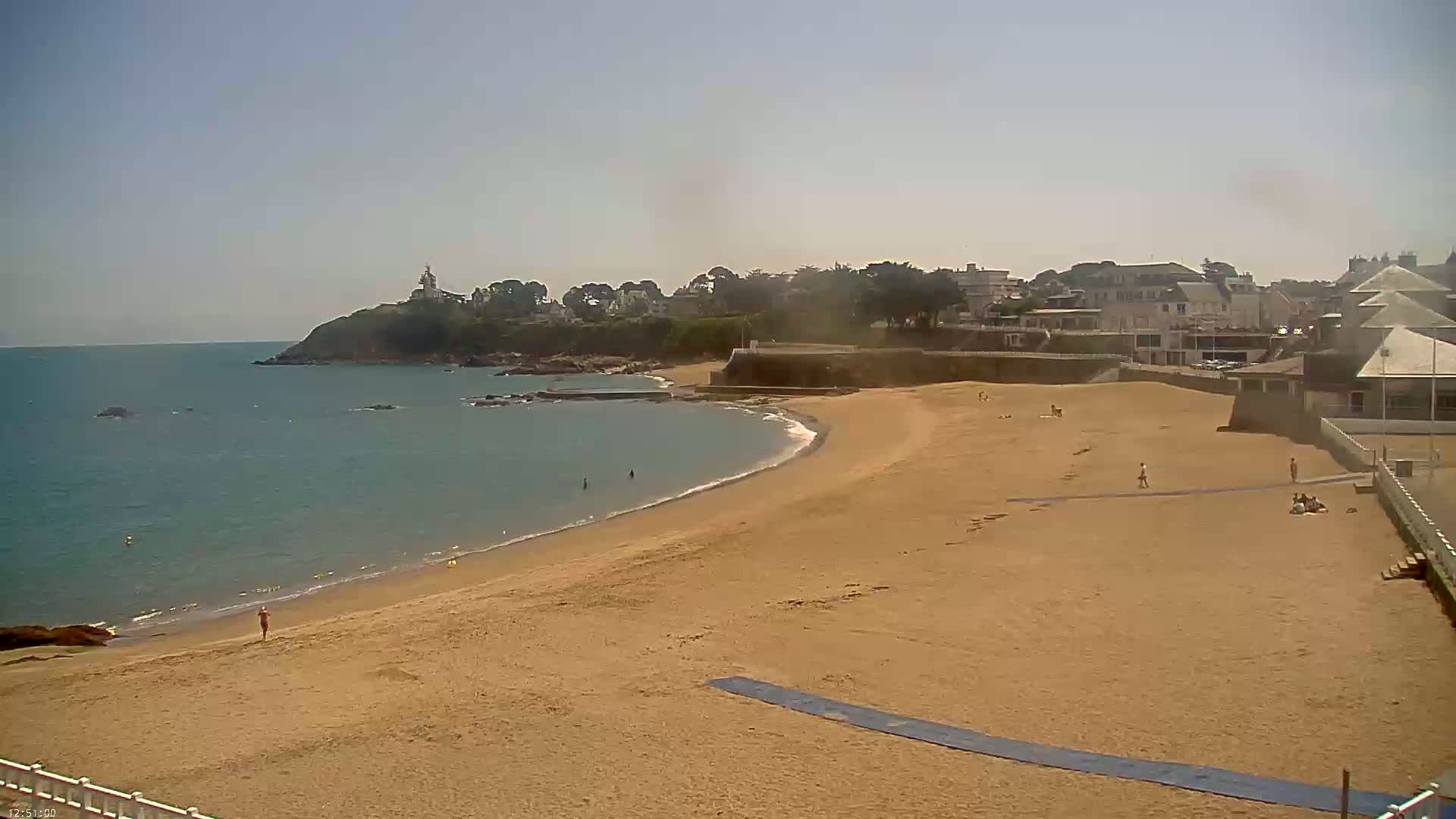 A mostly empty sandy beach curves around a calm, blue ocean, with a few people scattered on the sand and a small town visible in the background under a clear sky.