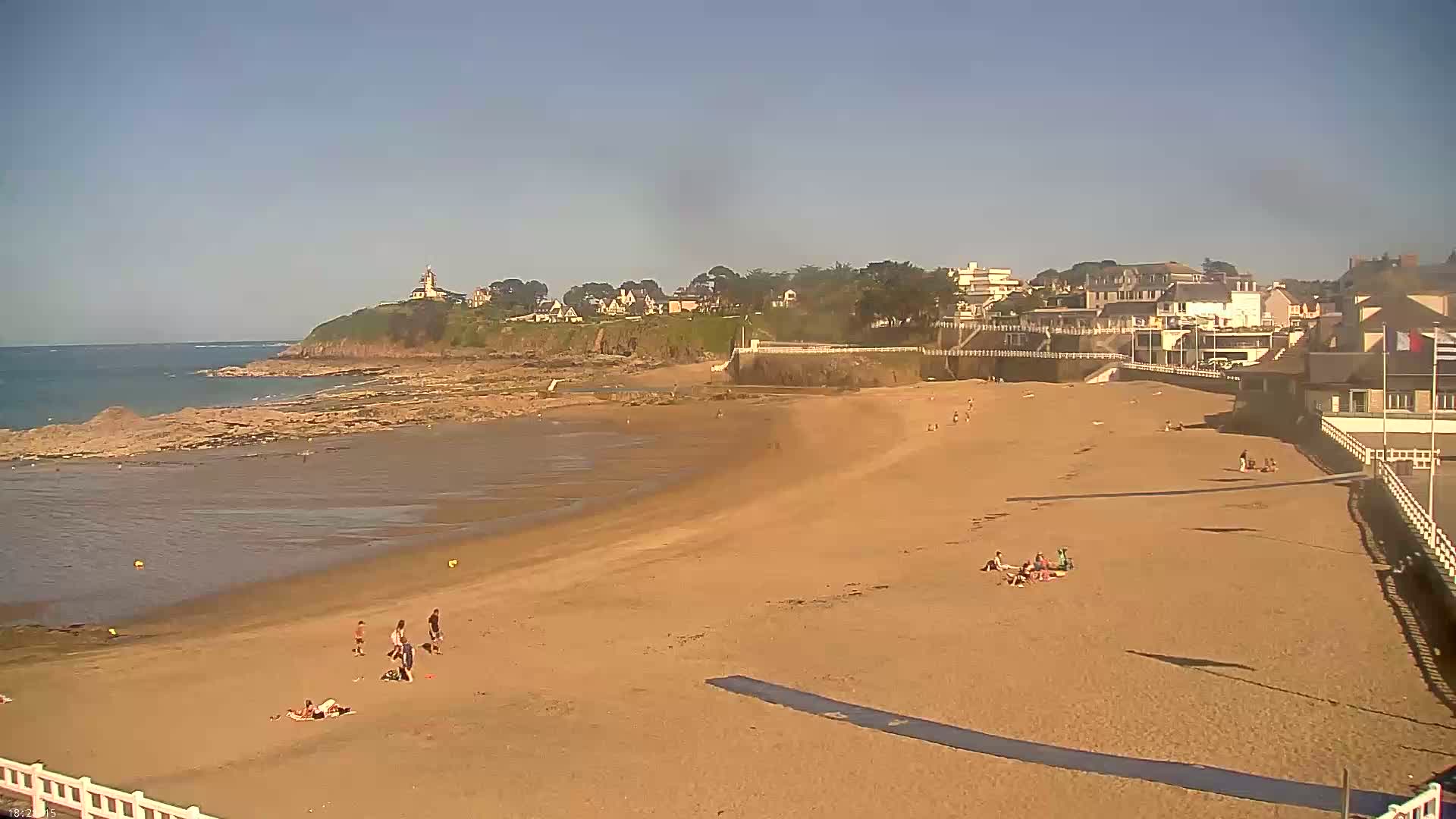 A sandy beach with a few people on it slopes down to a calm ocean under a clear, sunny sky, with buildings and a lighthouse visible on a cliff in the distance.