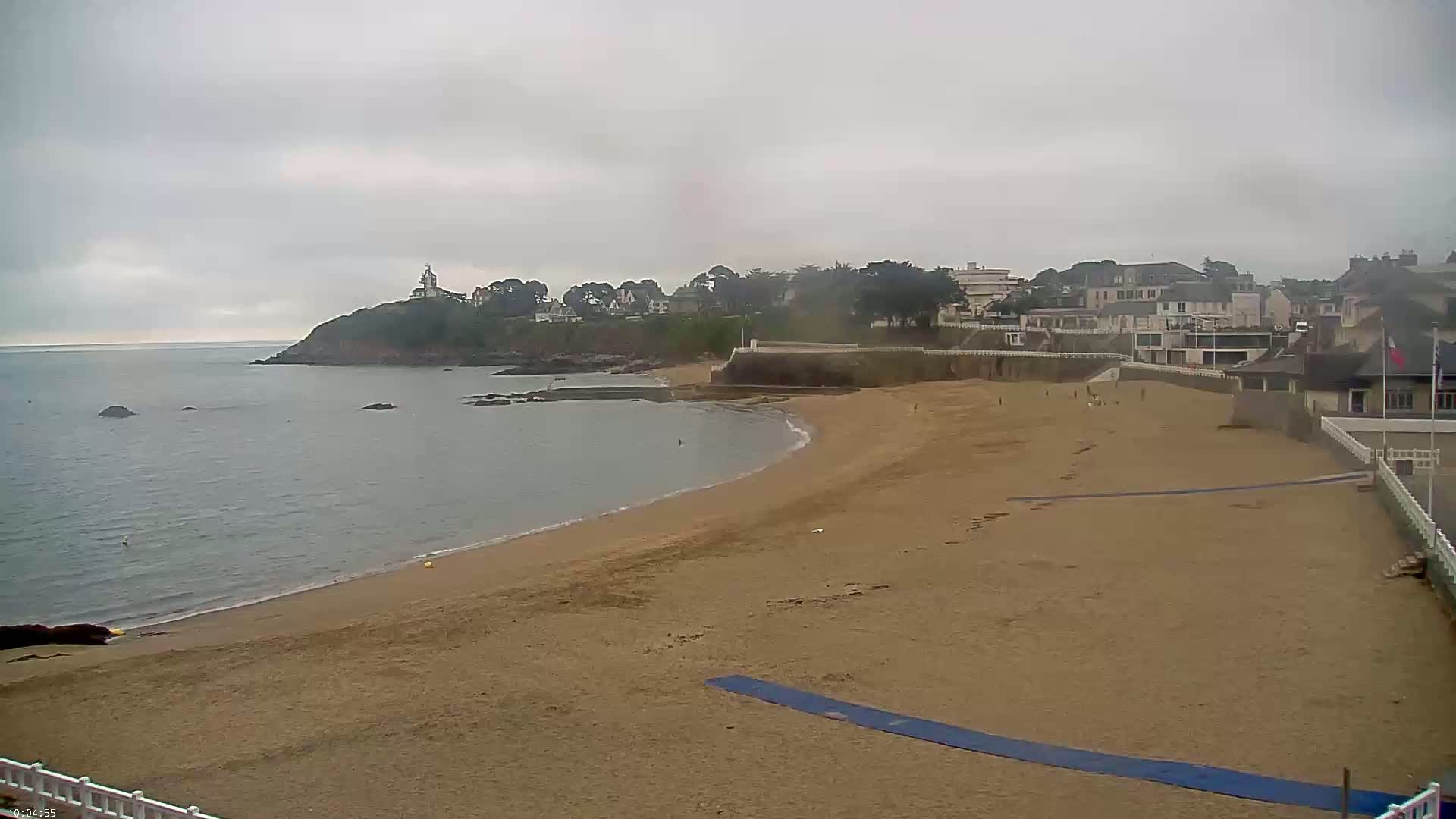 A sandy beach curves around a calm, gray sea under an overcast sky, with a small town and a lighthouse visible on a nearby headland.