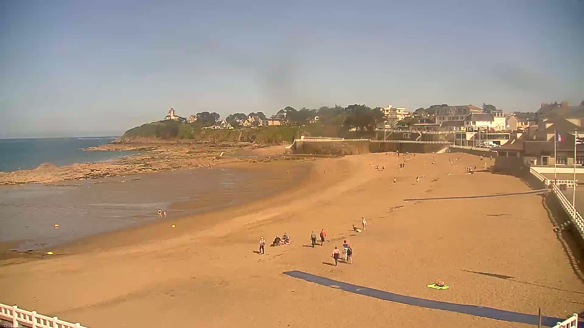 A sandy beach with a few people on it stretches out to a calm ocean under a clear, sunny sky, backed by buildings and green vegetation on a cliff.