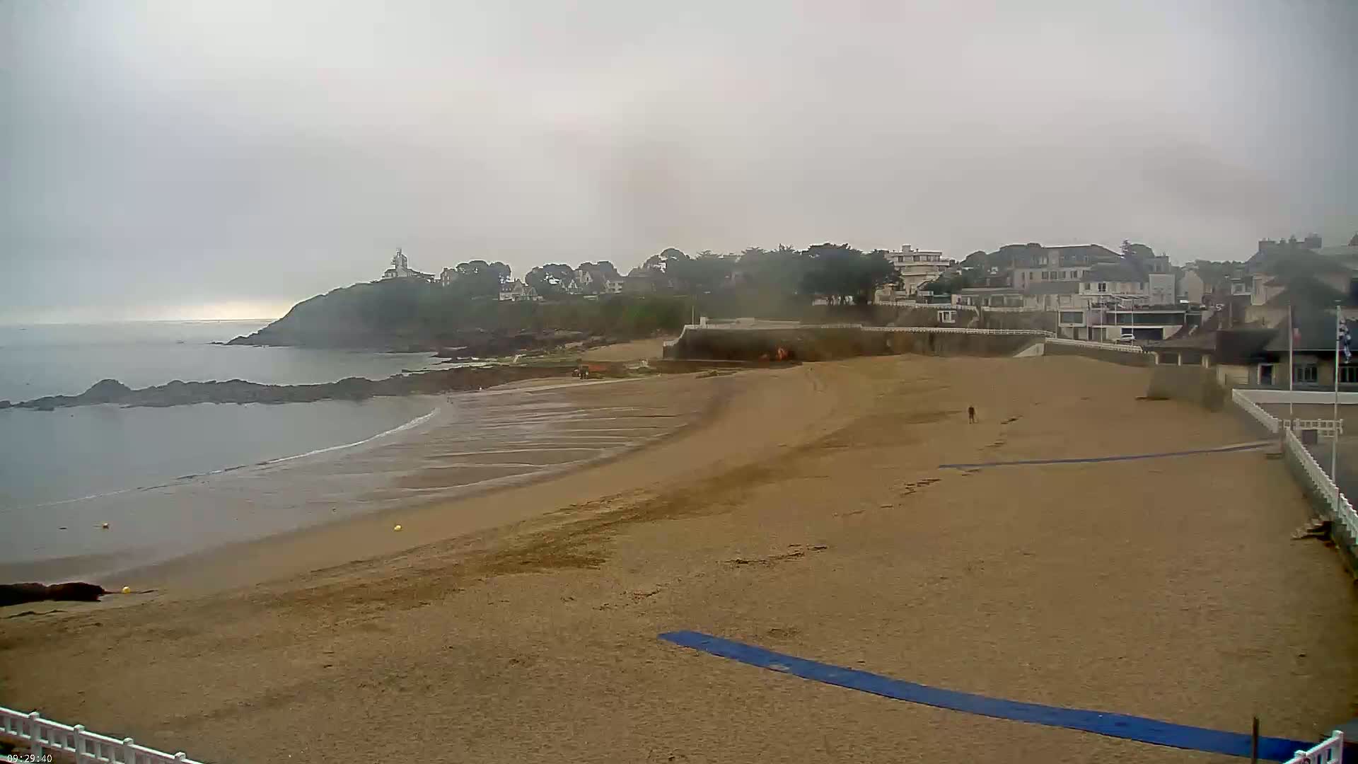 A sandy beach stretches out to a calm, grey sea under an overcast sky, with buildings and a small lighthouse visible in the distance on a rocky headland.