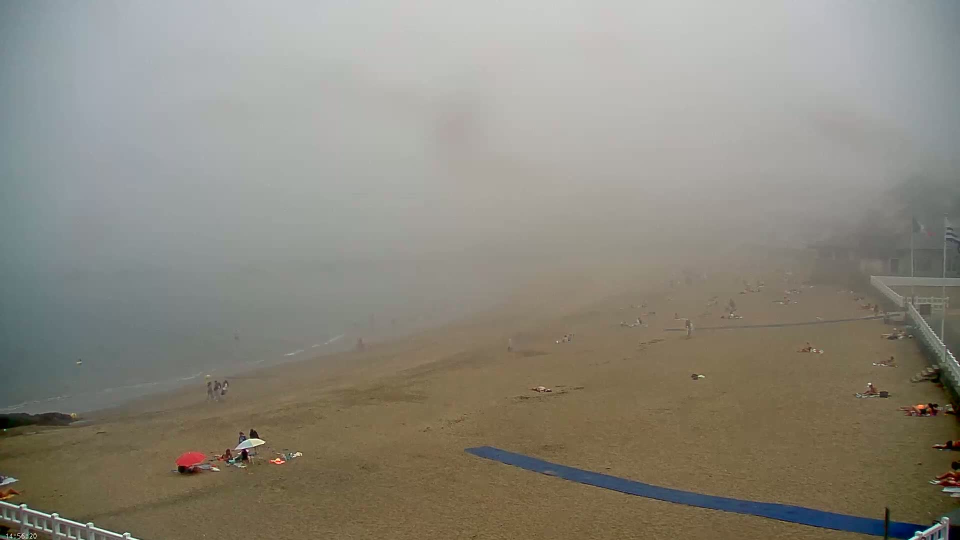 A foggy beach scene shows a sandy shore with scattered people sunbathing and relaxing near calm water.