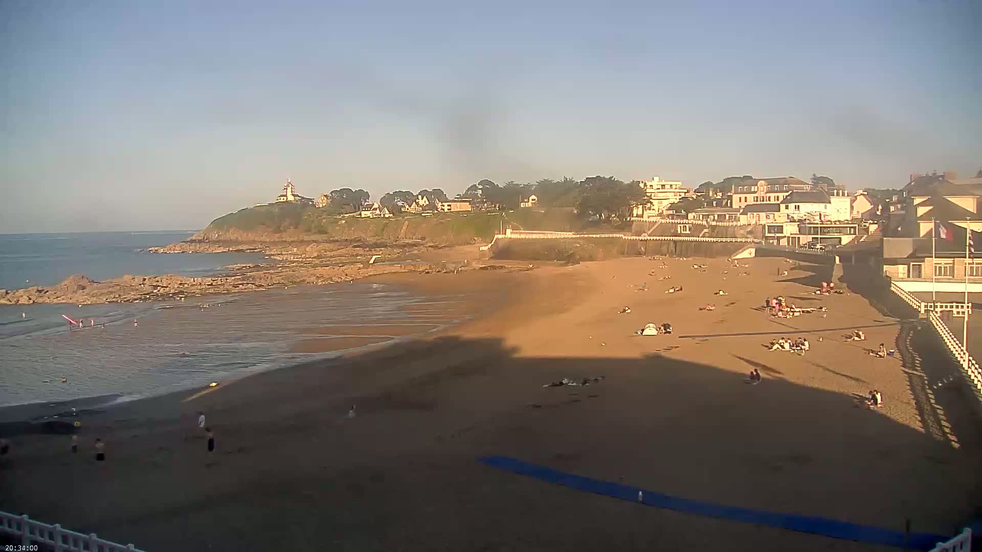 A sandy beach with scattered people basks in the late afternoon sun, adjacent to a calm ocean and buildings perched on a nearby cliff.