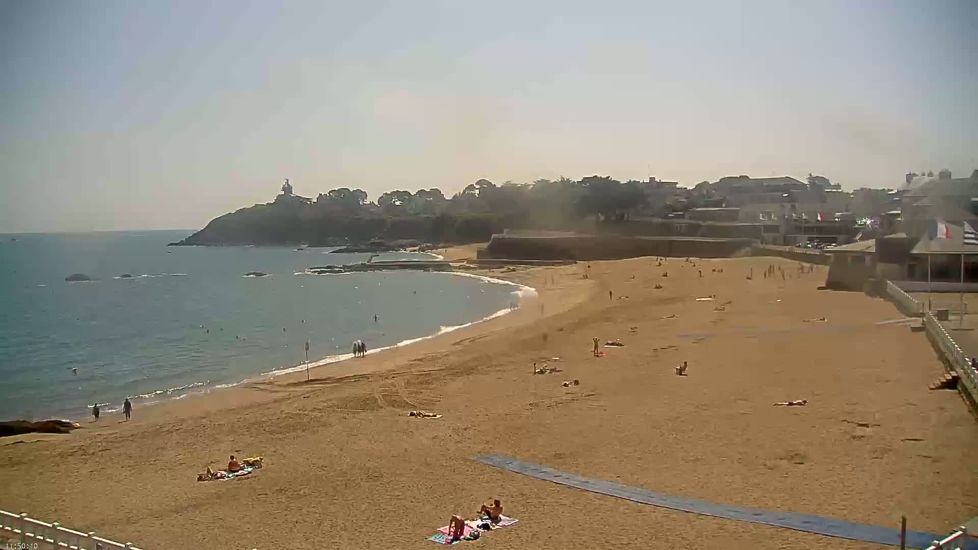A sandy beach with scattered sunbathers extends to a calm sea under a hazy sun.