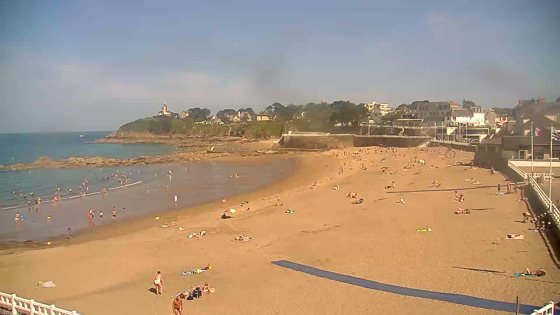 A sandy beach with many people swimming and sunbathing stretches along a calm ocean under a clear, sunny sky, backed by a town with buildings and a lighthouse on a cliff.