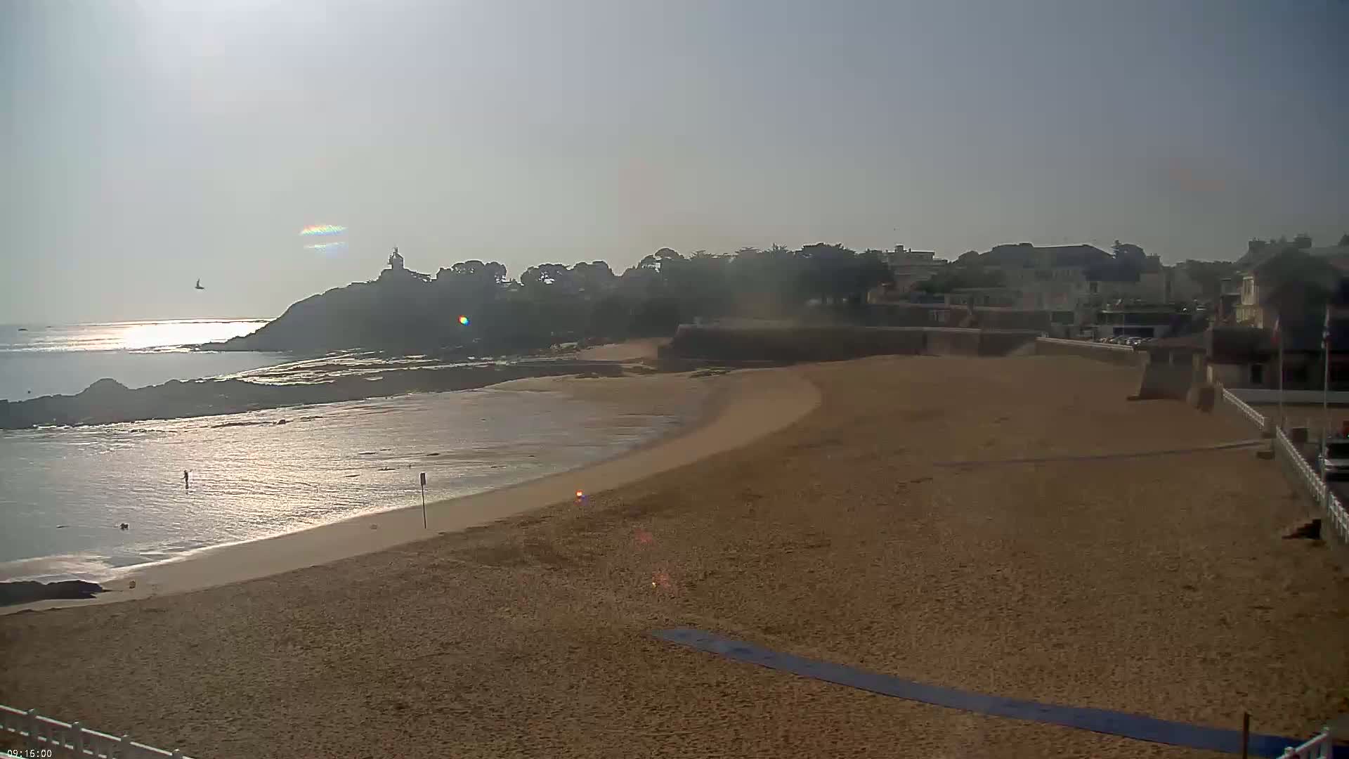 A sandy beach curves gently around a calm, sunlit ocean, with buildings and a small cliff in the background on a hazy day.