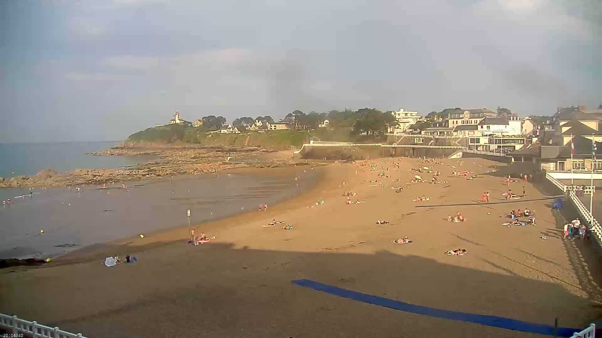 A sandy beach with many people relaxing and swimming, backed by buildings and a rocky coast under a cloudy sky.