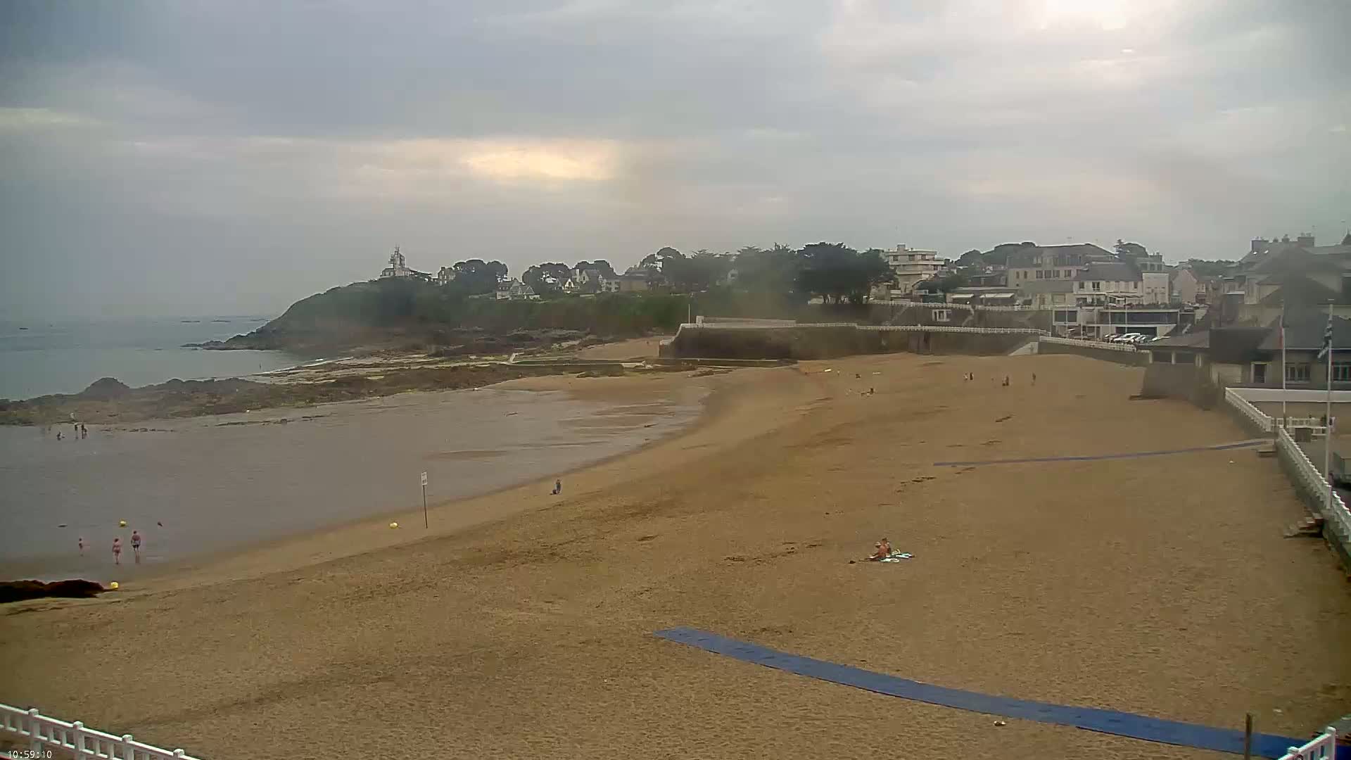 A sandy beach stretches towards a calm ocean under an overcast sky, with a few people scattered across the sand and a small town visible in the background.